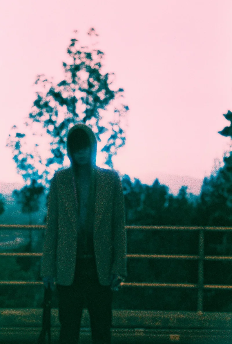 Soft-focus film frame of a hooded figure in a light blazer at a railing under a pink dusk sky