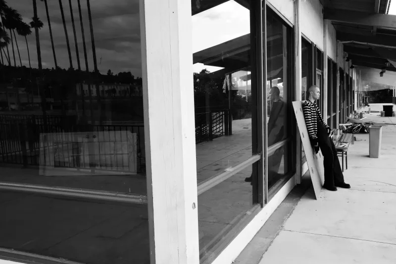 Black-and-white photo of a model in a striped shirt leaning by empty storefront windows reflecting palm trees