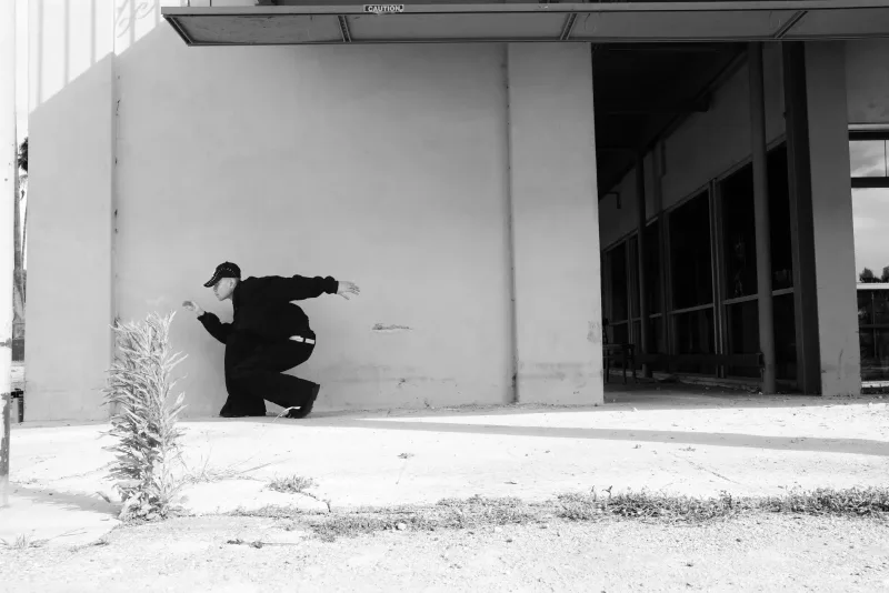 Black-and-white frame of a figure in dark tailoring bracing against the white wall of an empty building