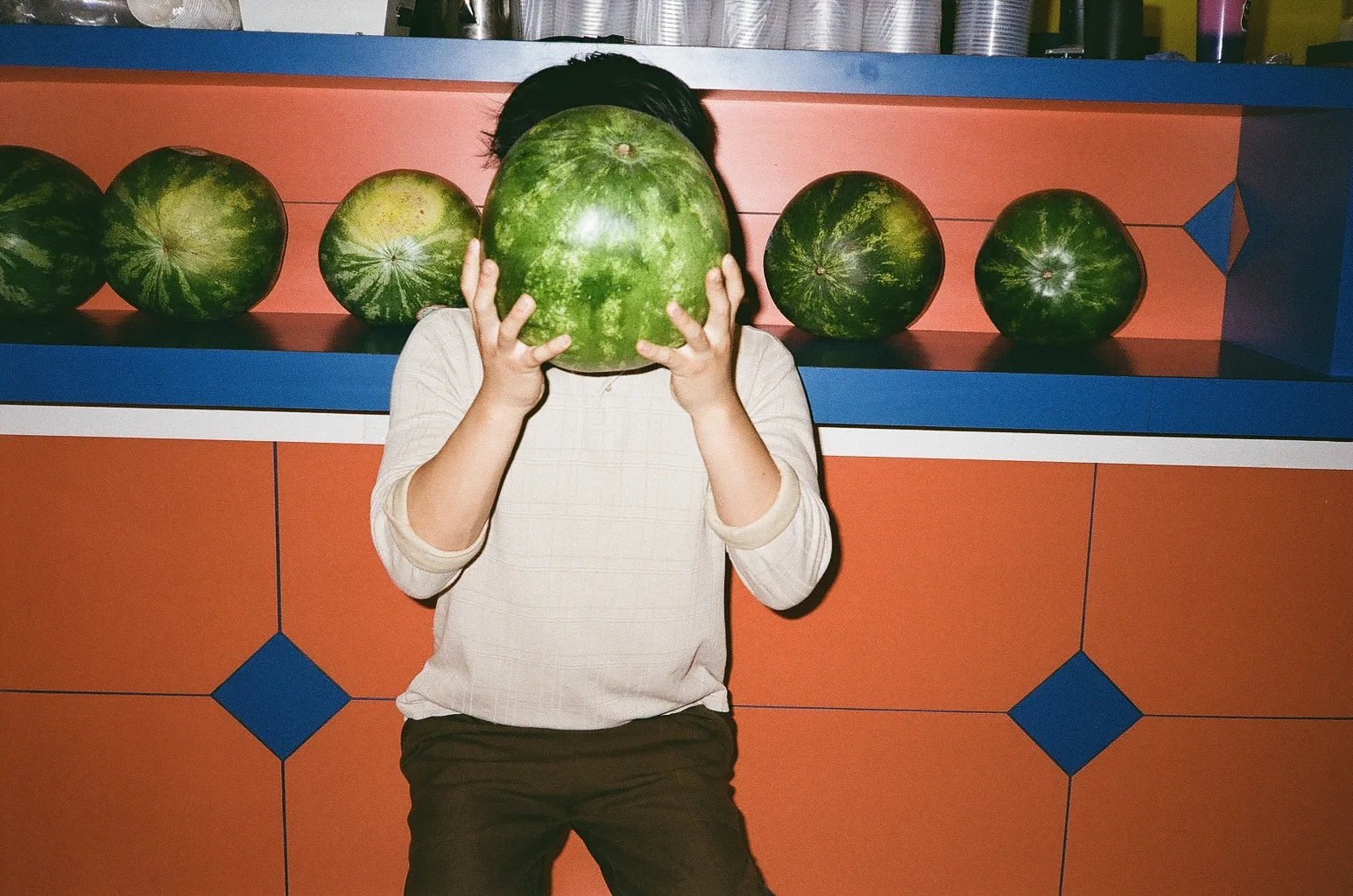 Person holding a watermelon in front of their face beside a market shelf of melons