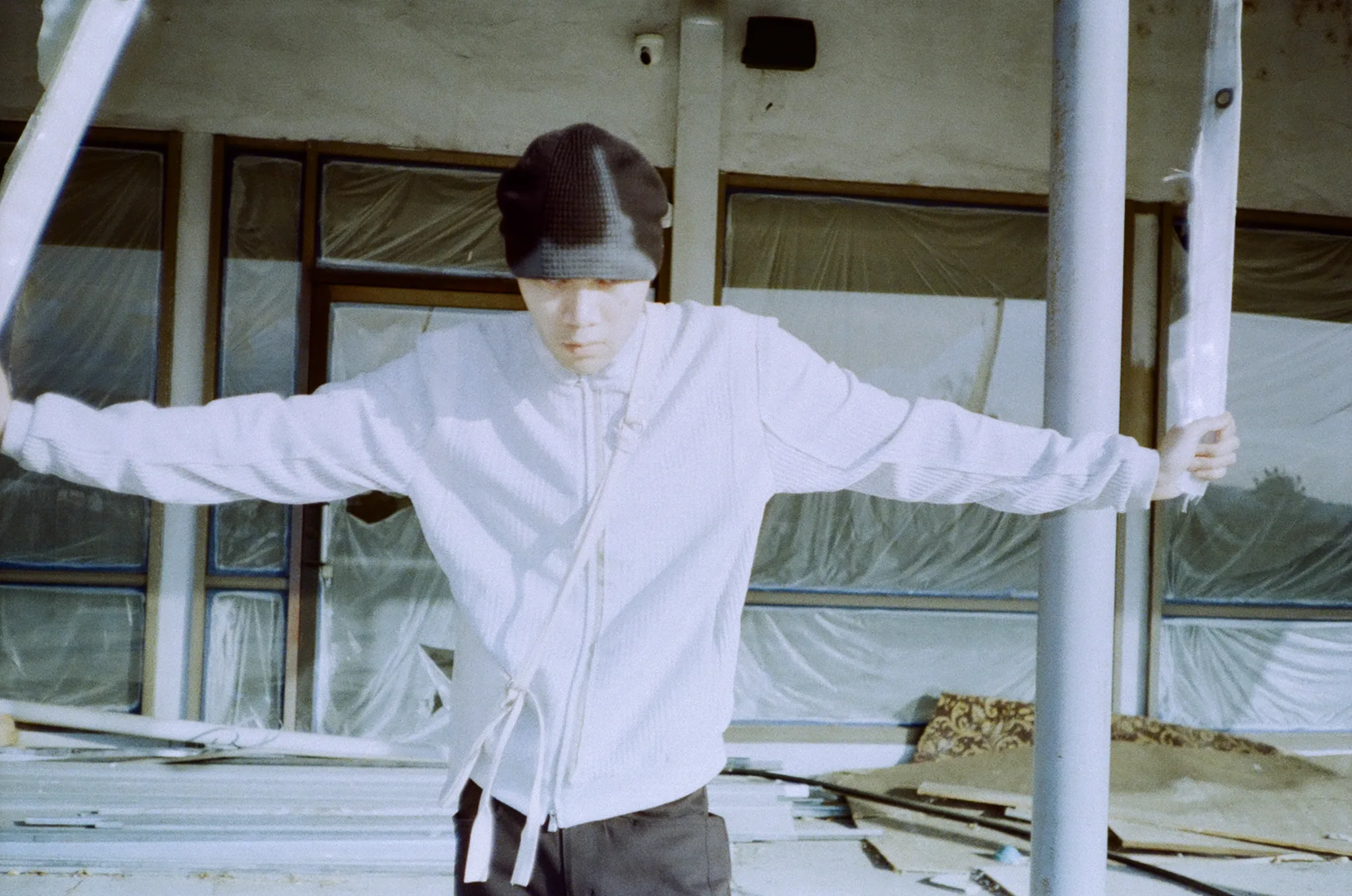 Model in a white jacket and beanie holding two poles in front of a building wrapped in plastic sheeting, on film