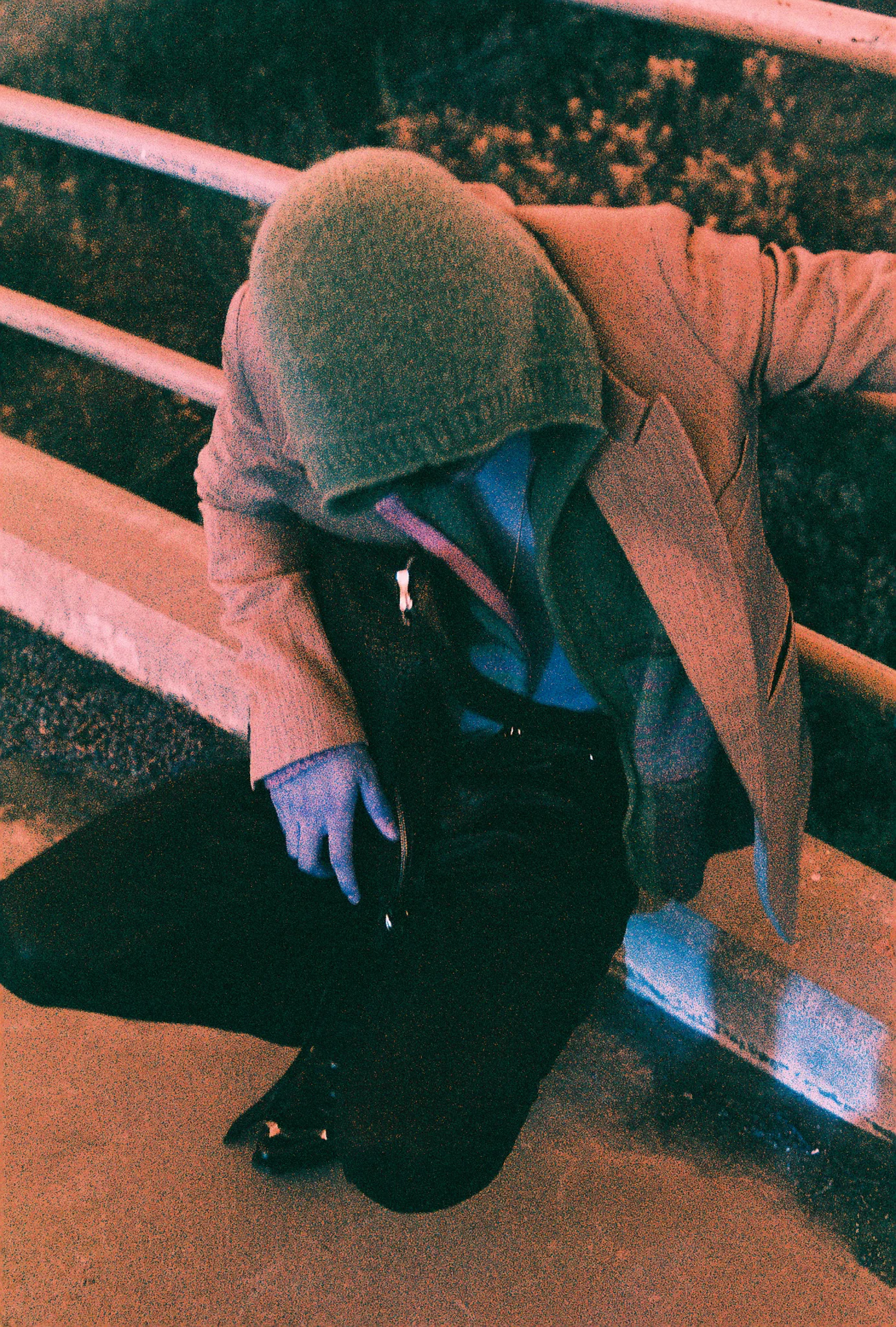 Figure in a knit hood and tan coat crouched against a railing at night, shot on film