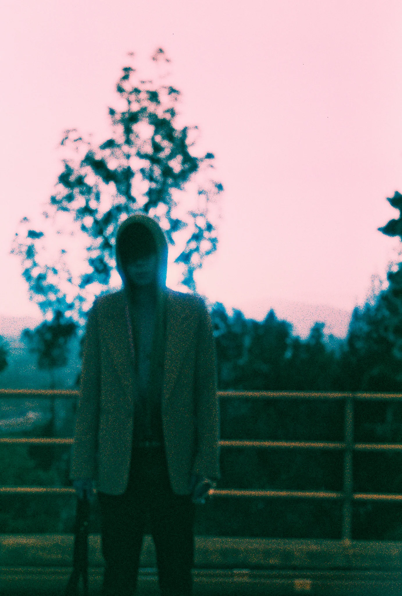 Soft-focus film frame of a hooded figure in a light blazer at a railing under a pink dusk sky