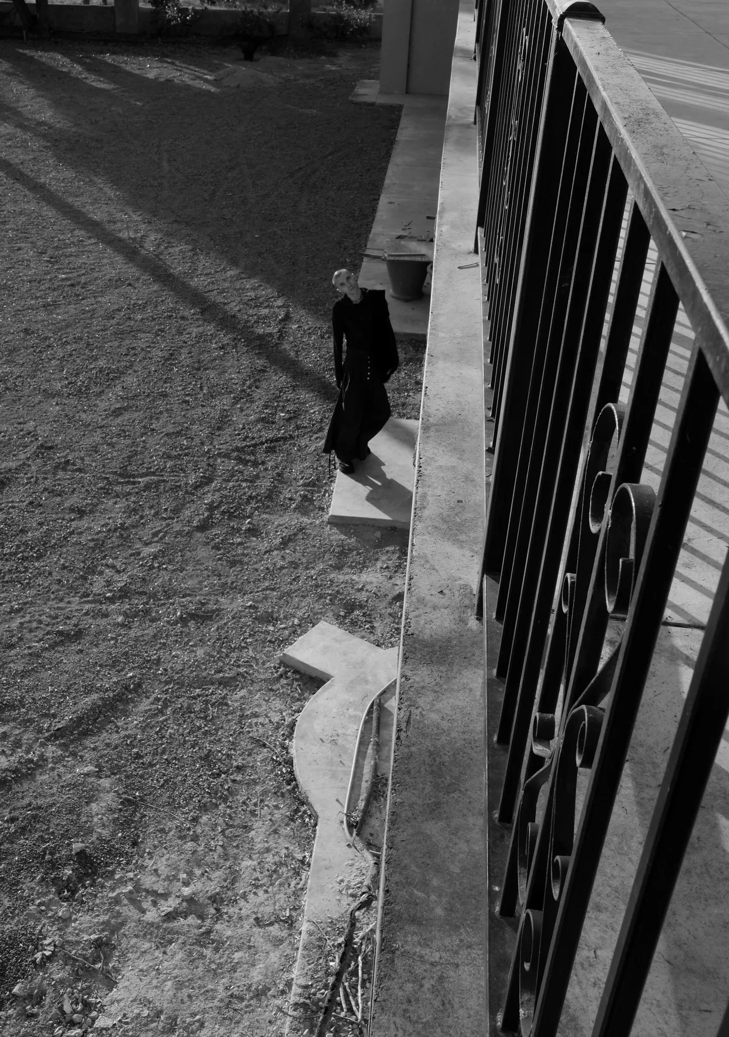 Black-and-white high-angle view past an iron railing of a figure in a long dark coat on gravel below