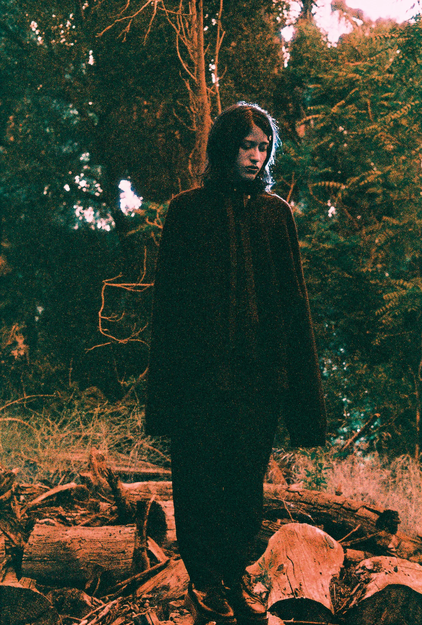 Model in a long dark coat standing in woodland over felled logs at dusk, on film