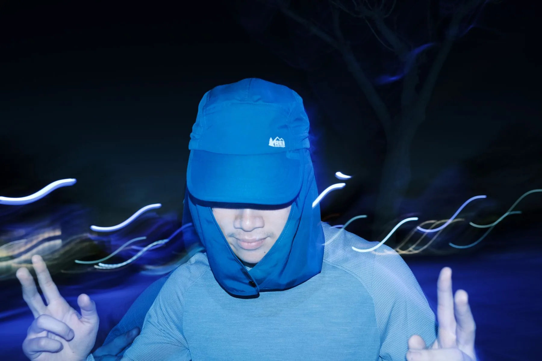 Person in a blue flap cap flashing peace signs among light trails at night