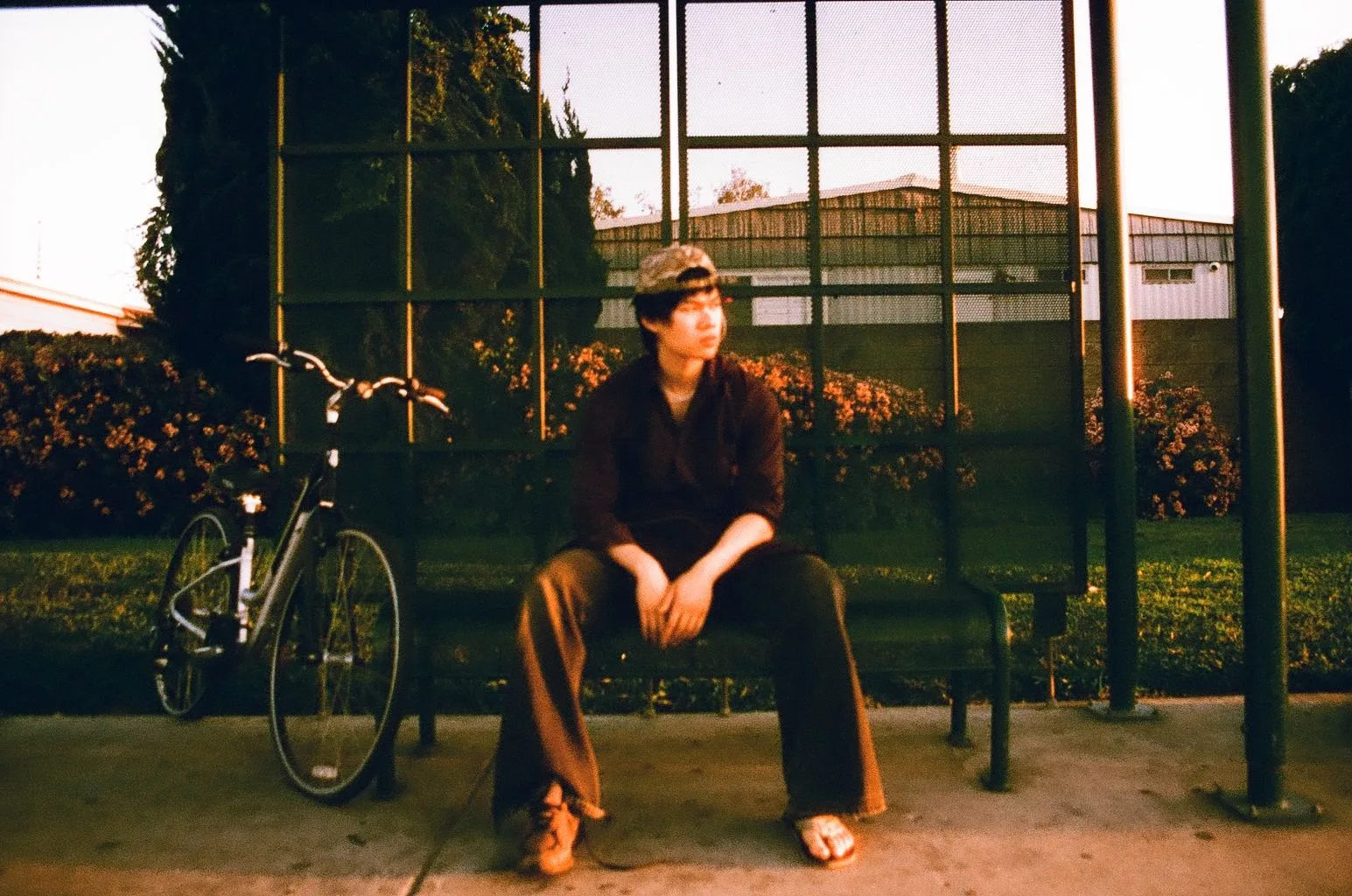 Person in a cap seated on a bench beside a bicycle at golden hour, on film