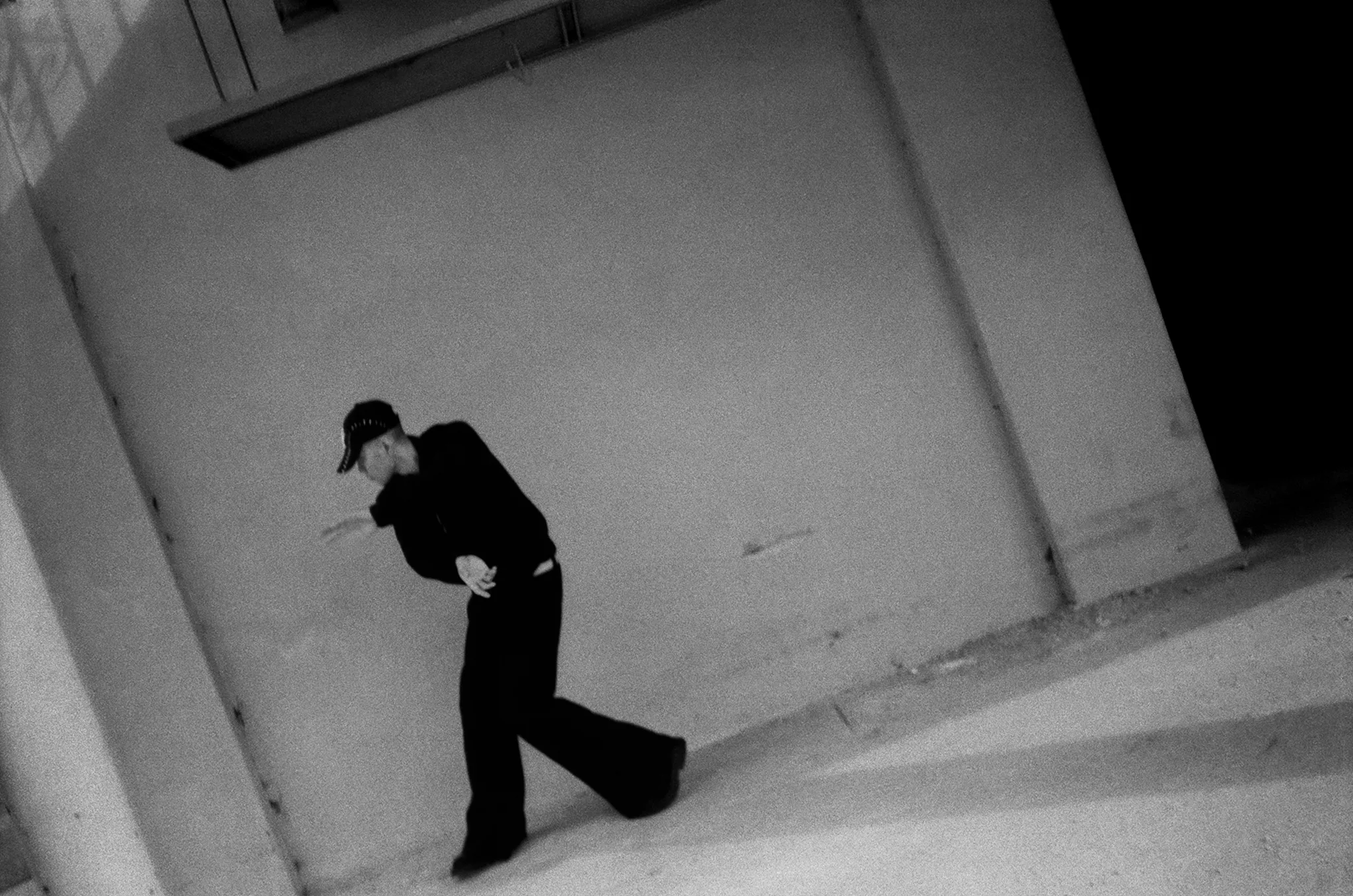 Black-and-white frame of a figure in black mid-swing in an empty concrete lot
