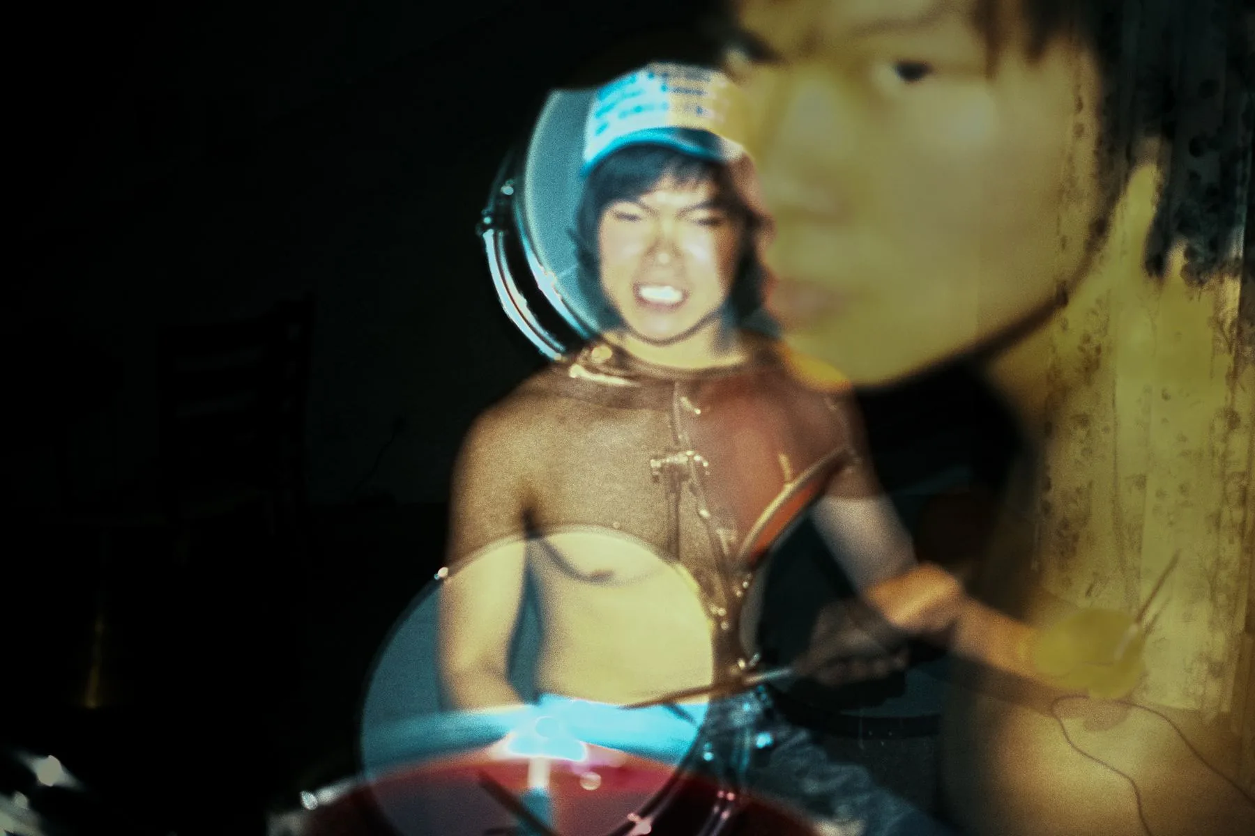 Double exposure of a shirtless drummer in a trucker cap mid-performance