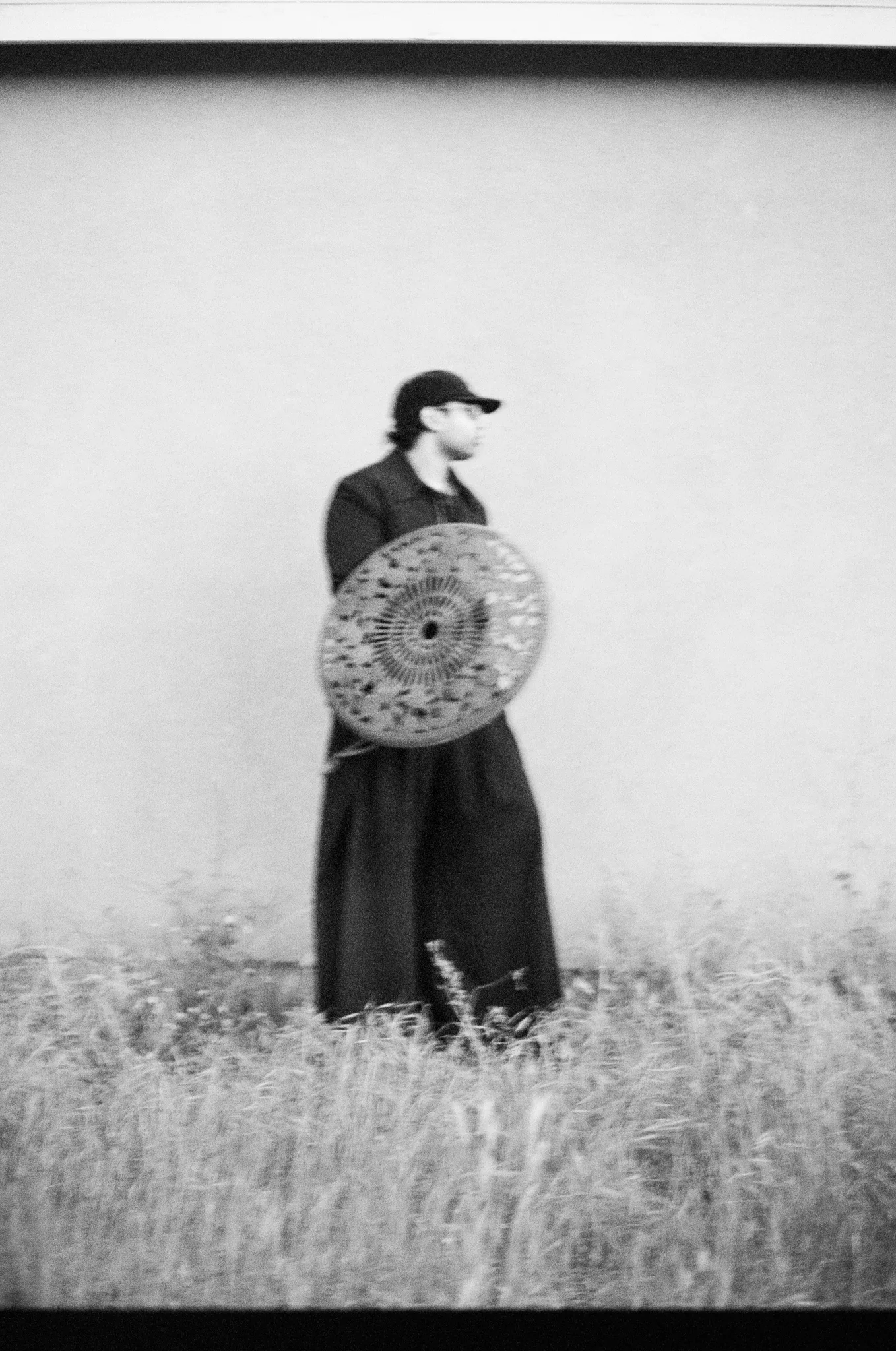 Man in black holding a mosaic disc like a shield in tall grass, on film