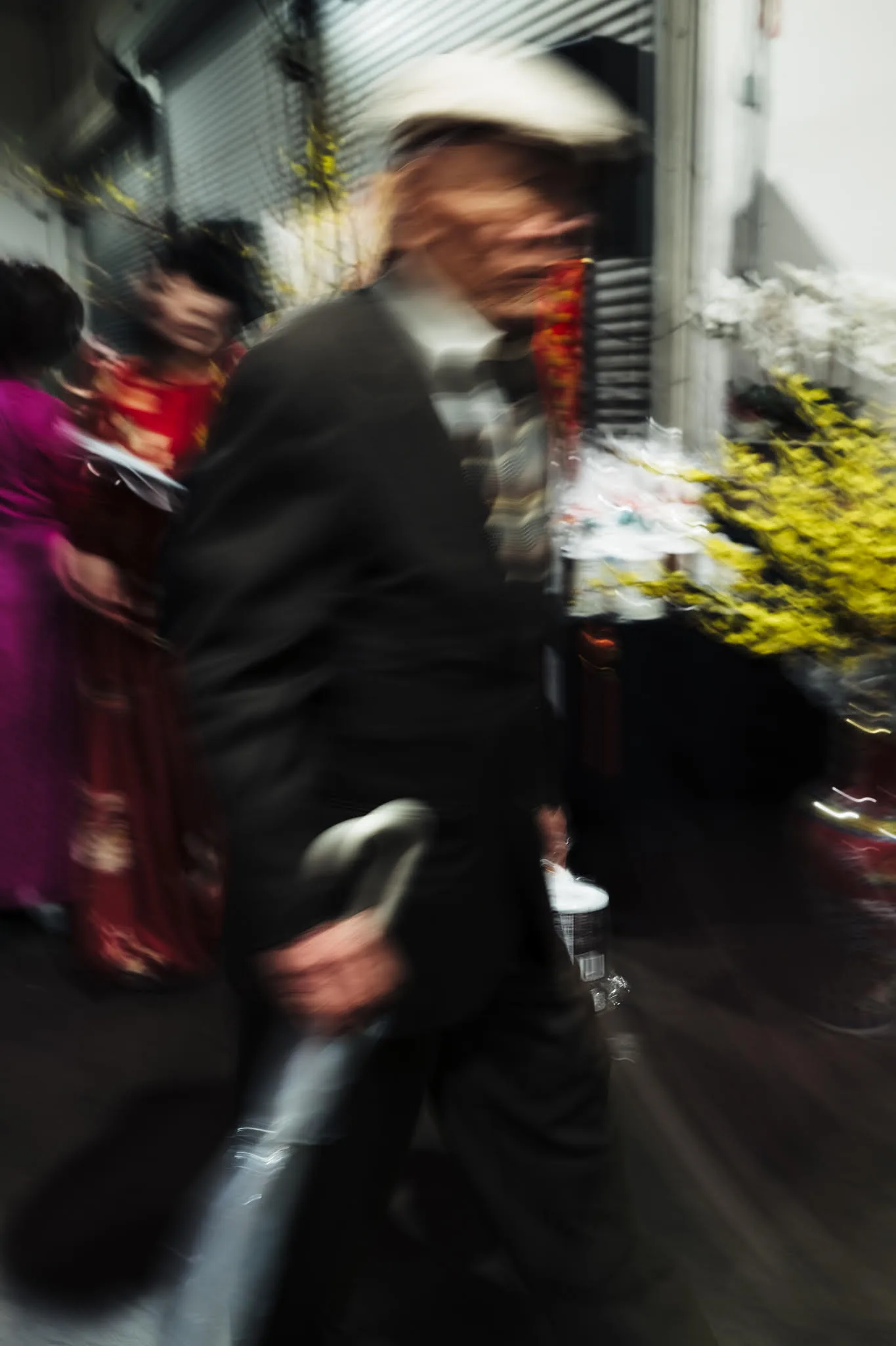 Motion-blurred elder in a white cap walking with a cane through a Tết flower market