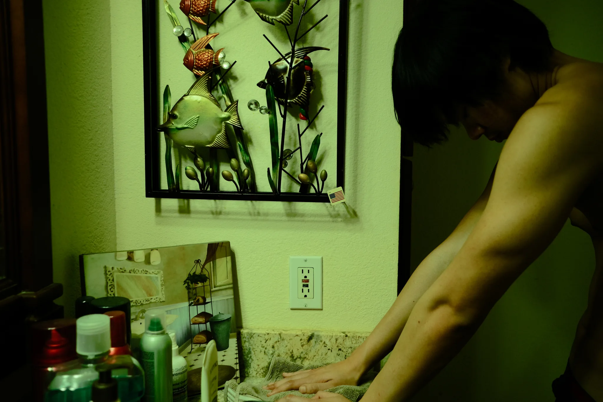 Figure washing up at a sink beneath a metal fish wall hanging in a green-lit bathroom