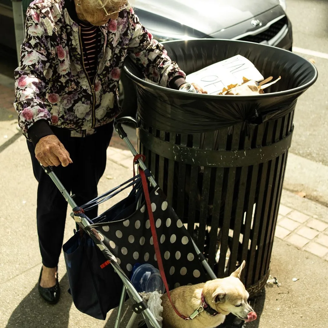 Elderly woman in a floral jacket with a walker and small dog beside a street trash can