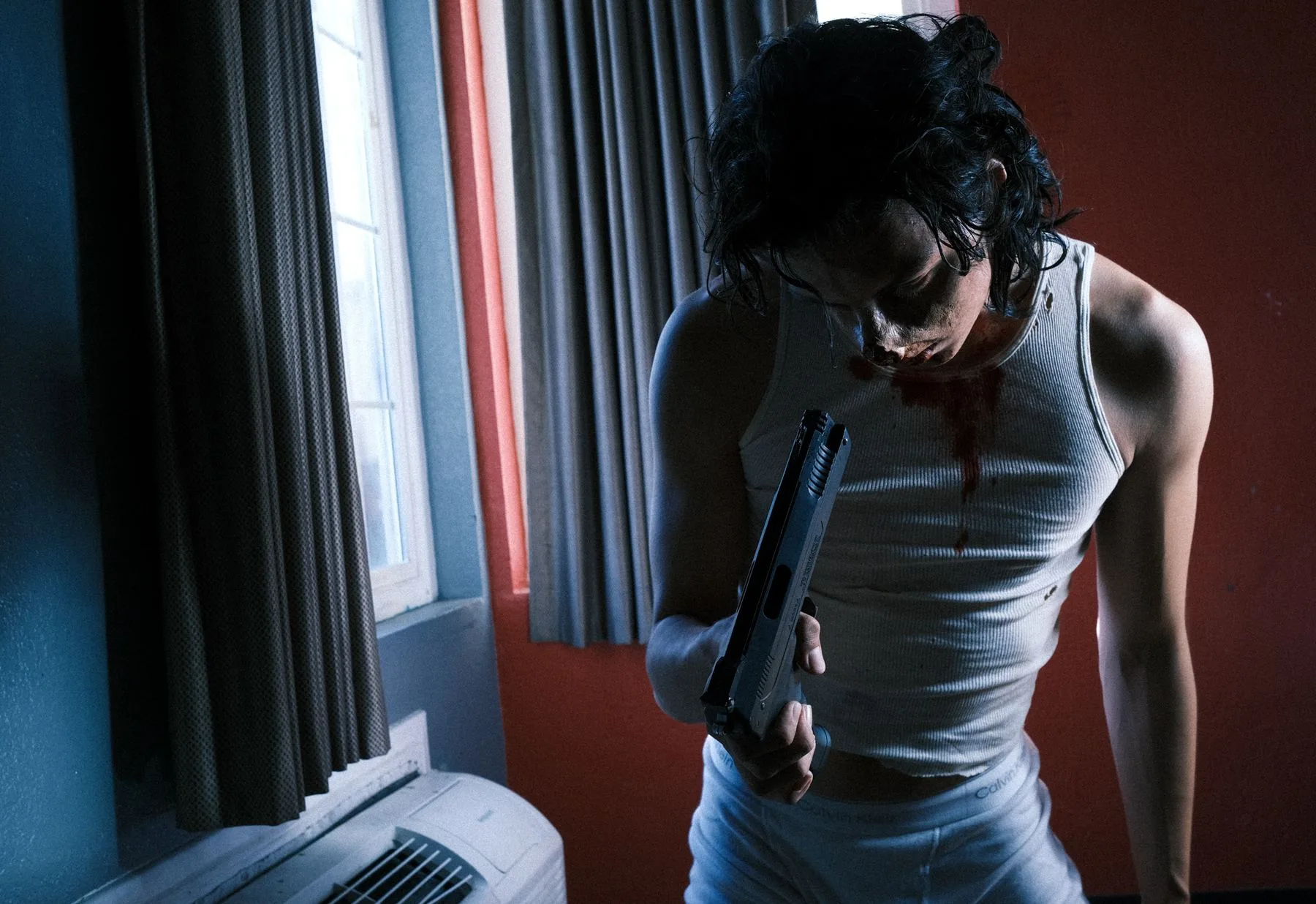 Staged motel-room scene in red and blue light, a figure in a white tank holding a prop pistol
