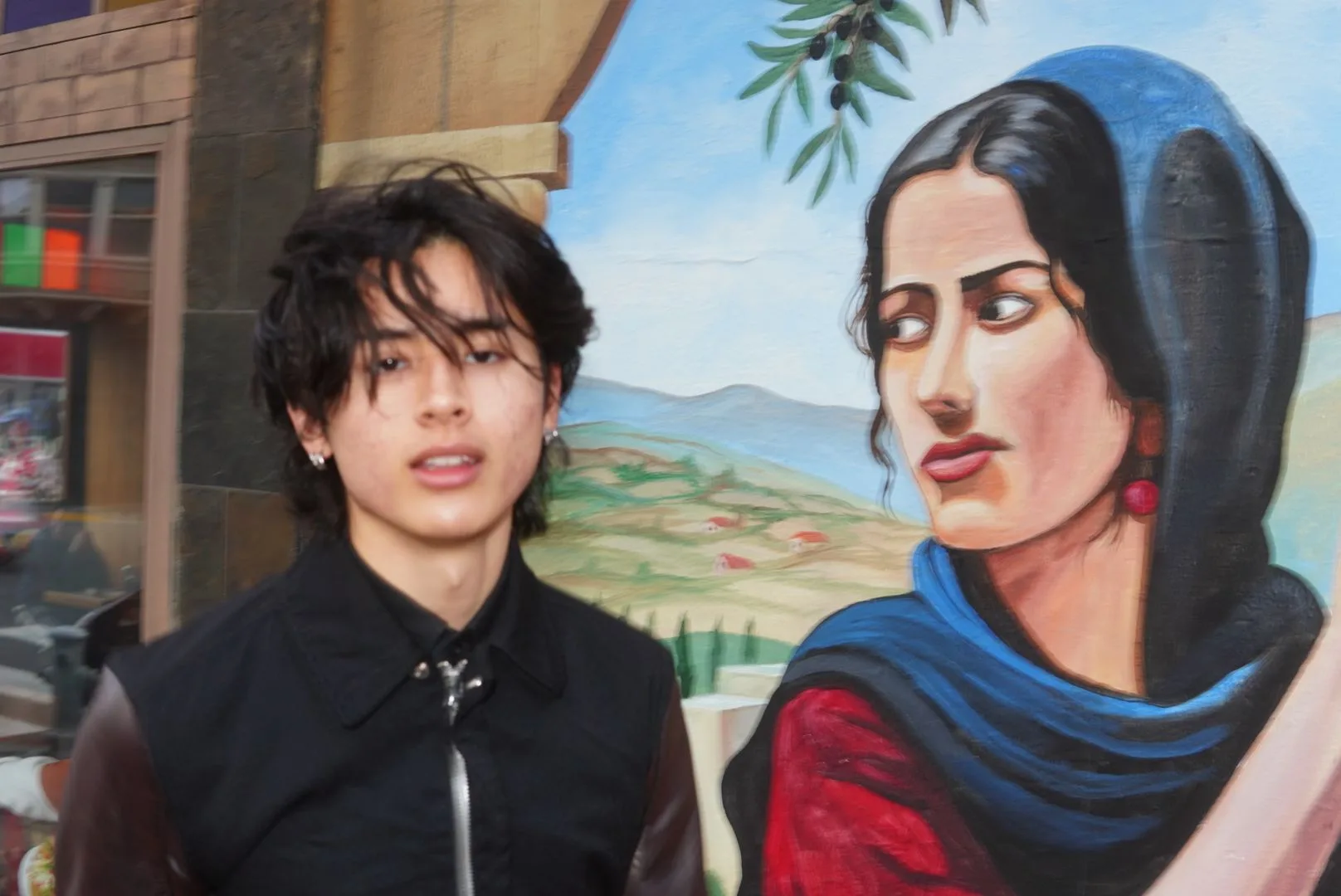 Young man in black beside a painted mural woman, matching her sidelong glance
