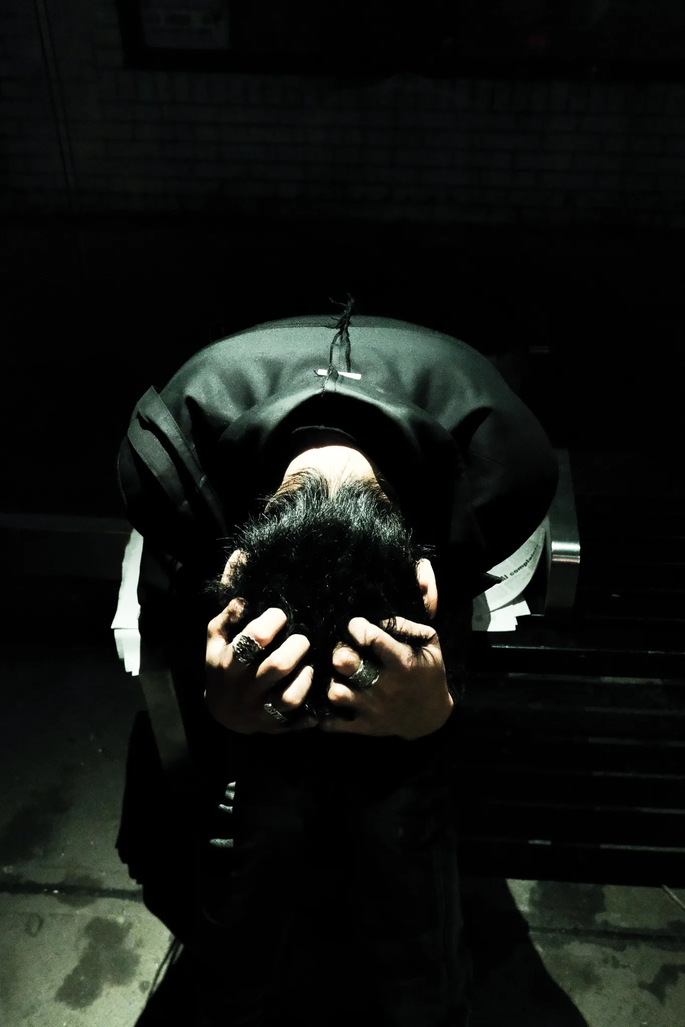Figure in black bowed forward, ringed hands gripping his hair in low light