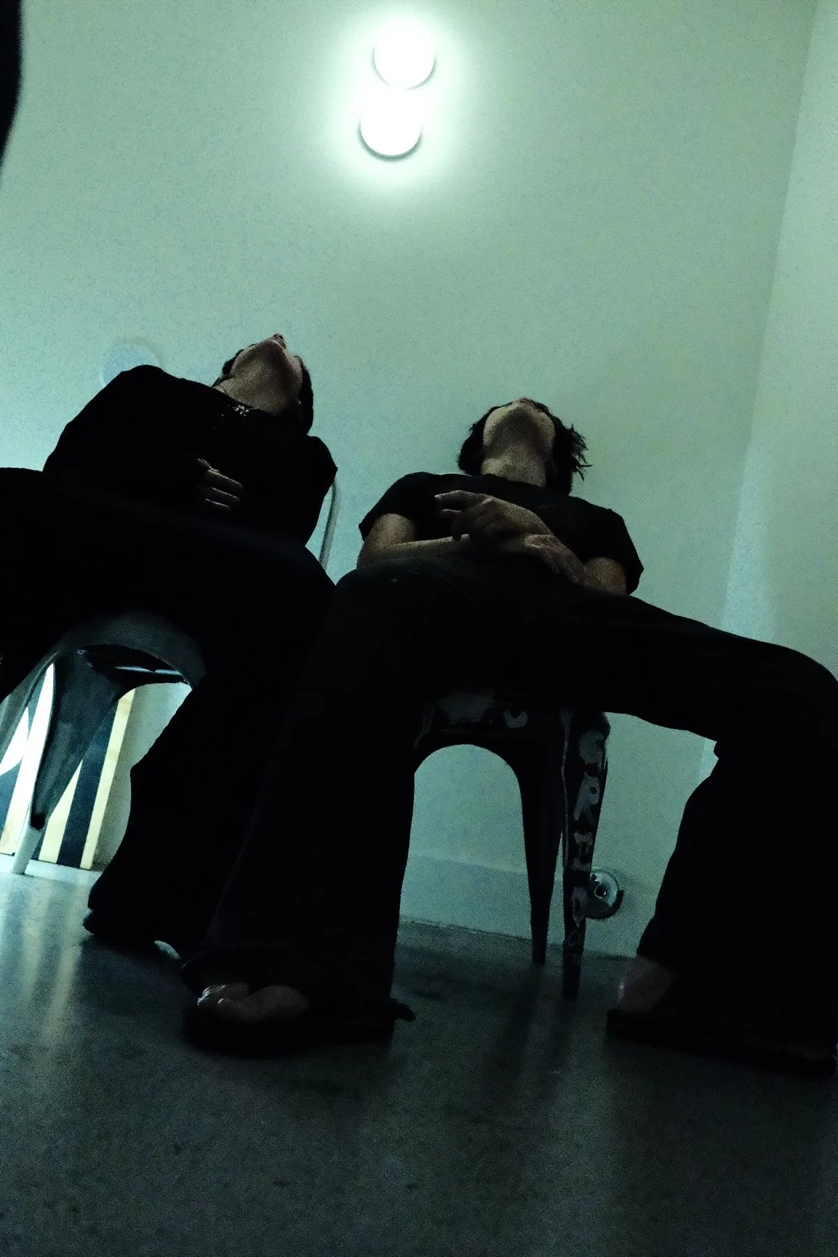 Two figures slumped asleep in chairs beneath a fluorescent lamp, teal-toned
