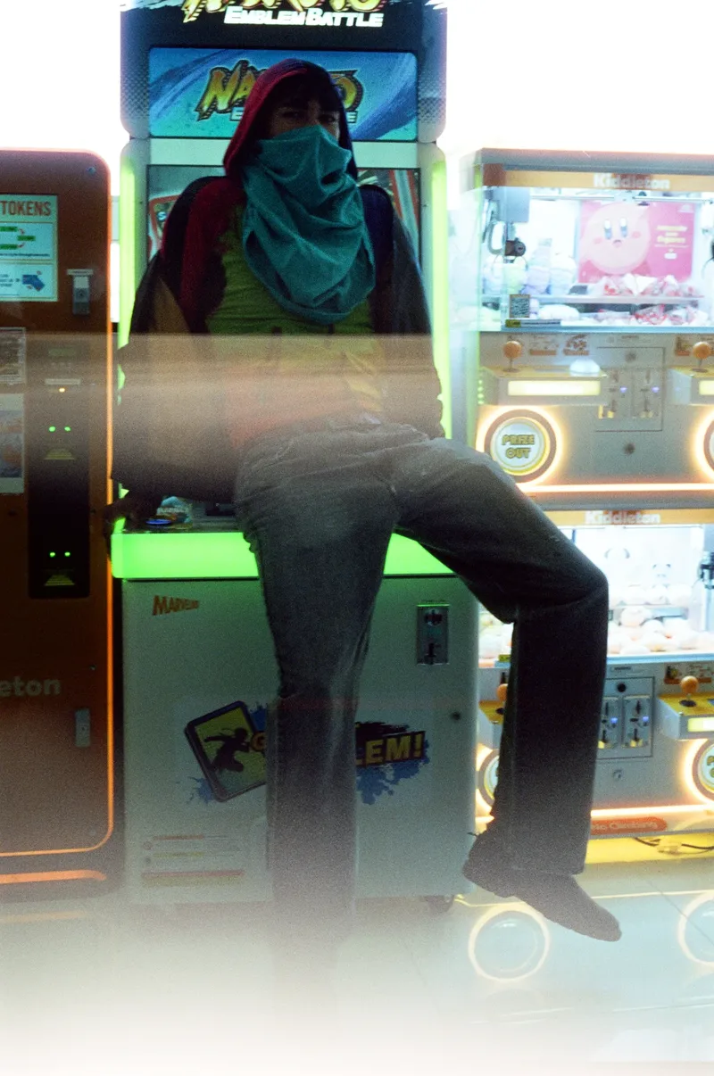 Figure with a teal scarf over their face seated on an arcade cabinet between claw machines, shot on film