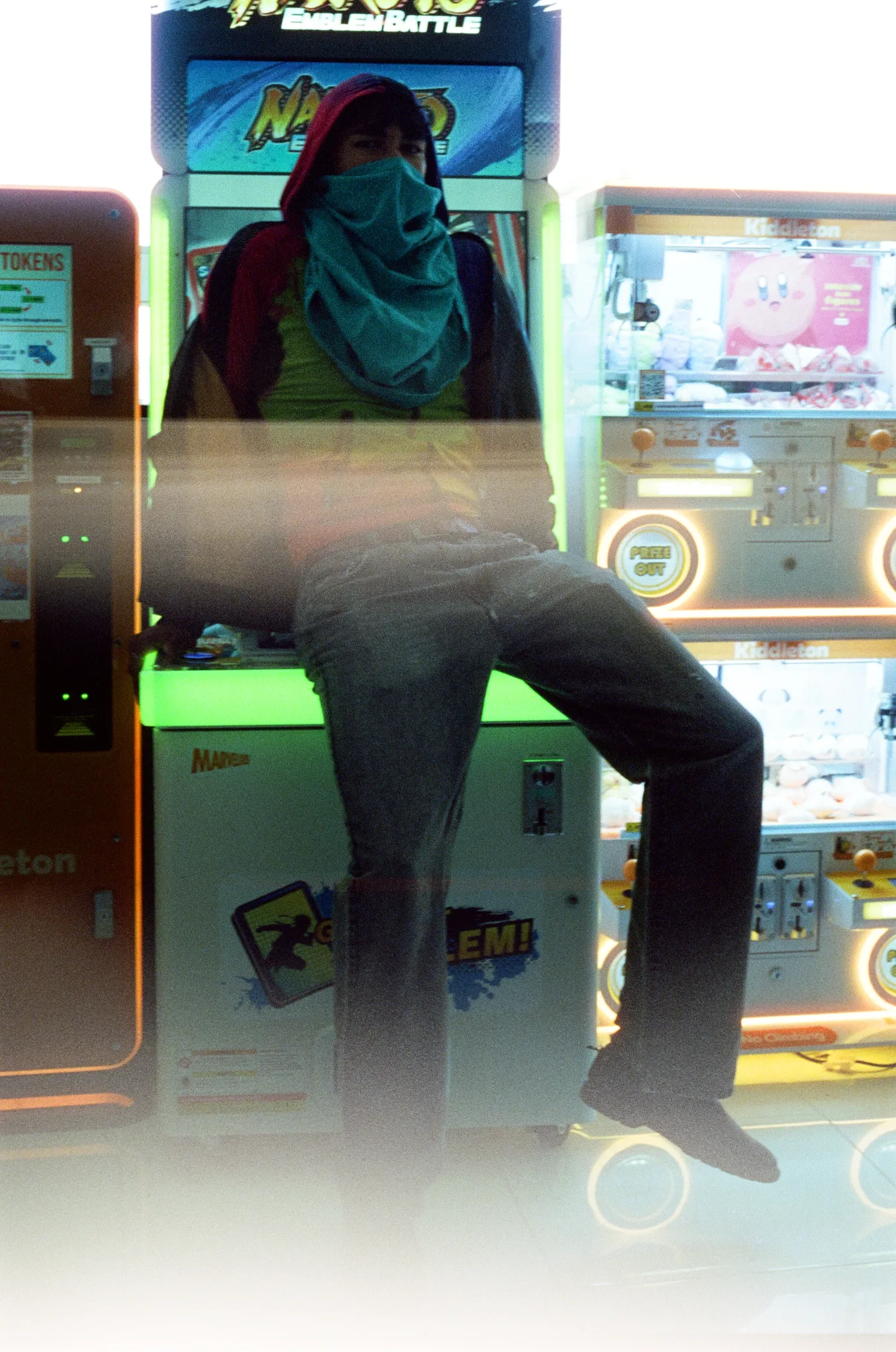 Figure with a teal scarf over their face seated on an arcade cabinet between claw machines, shot on film