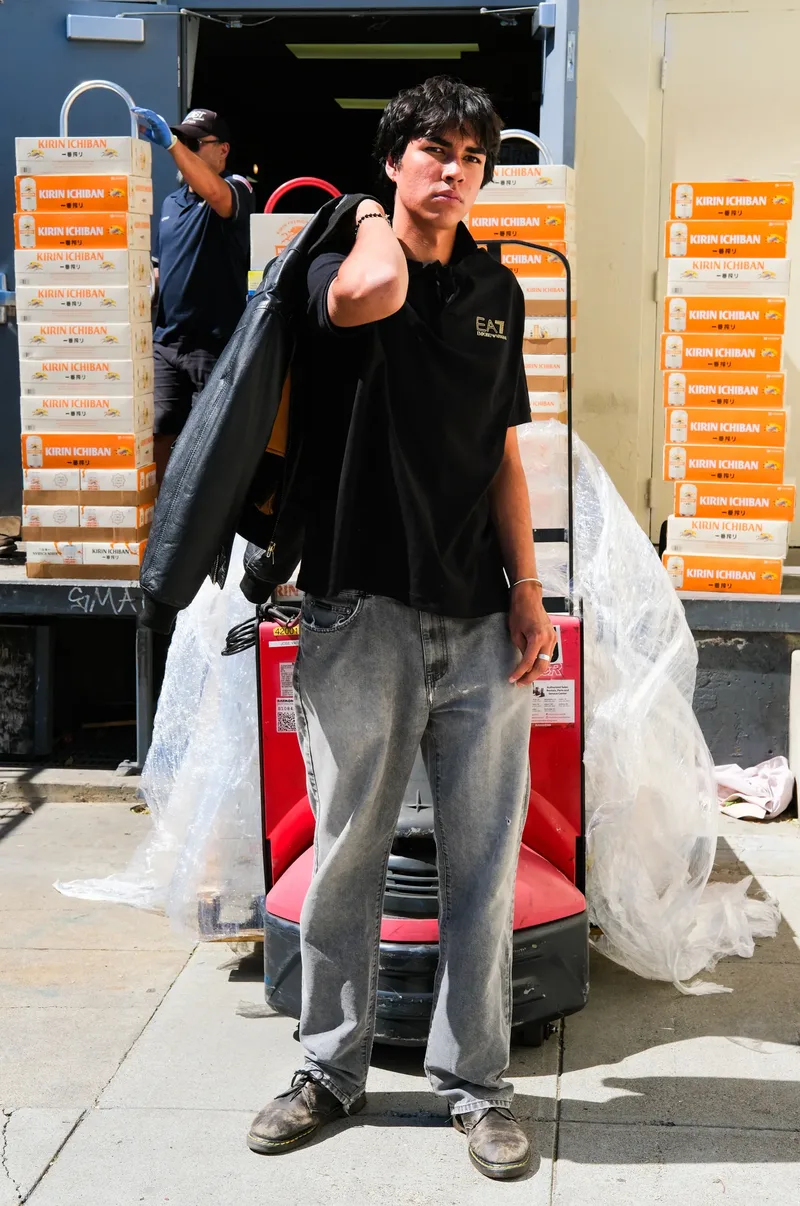 Model in a black polo and grey jeans holding a garment bag in front of a forklift and stacked beer crates