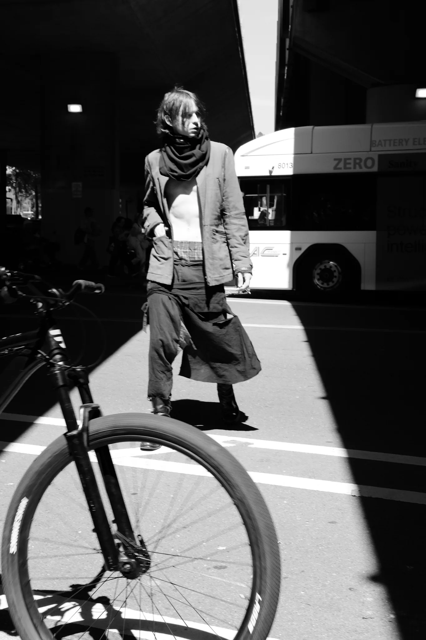 Black-and-white street photo of a model in a scarf and layered vintage outfit passing a city bus, bicycle wheel in the foreground
