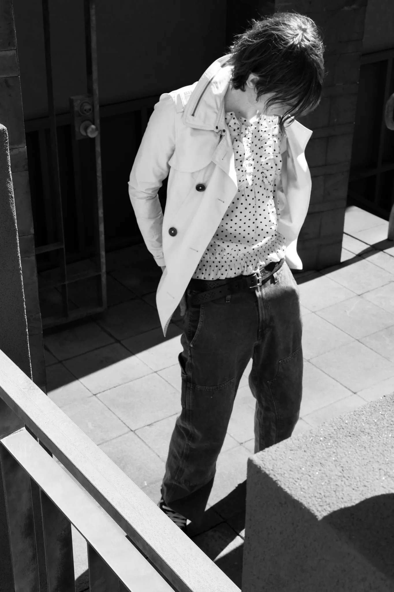 Black-and-white portrait of a model in a light trench and polka-dot shirt looking down on a tiled walkway