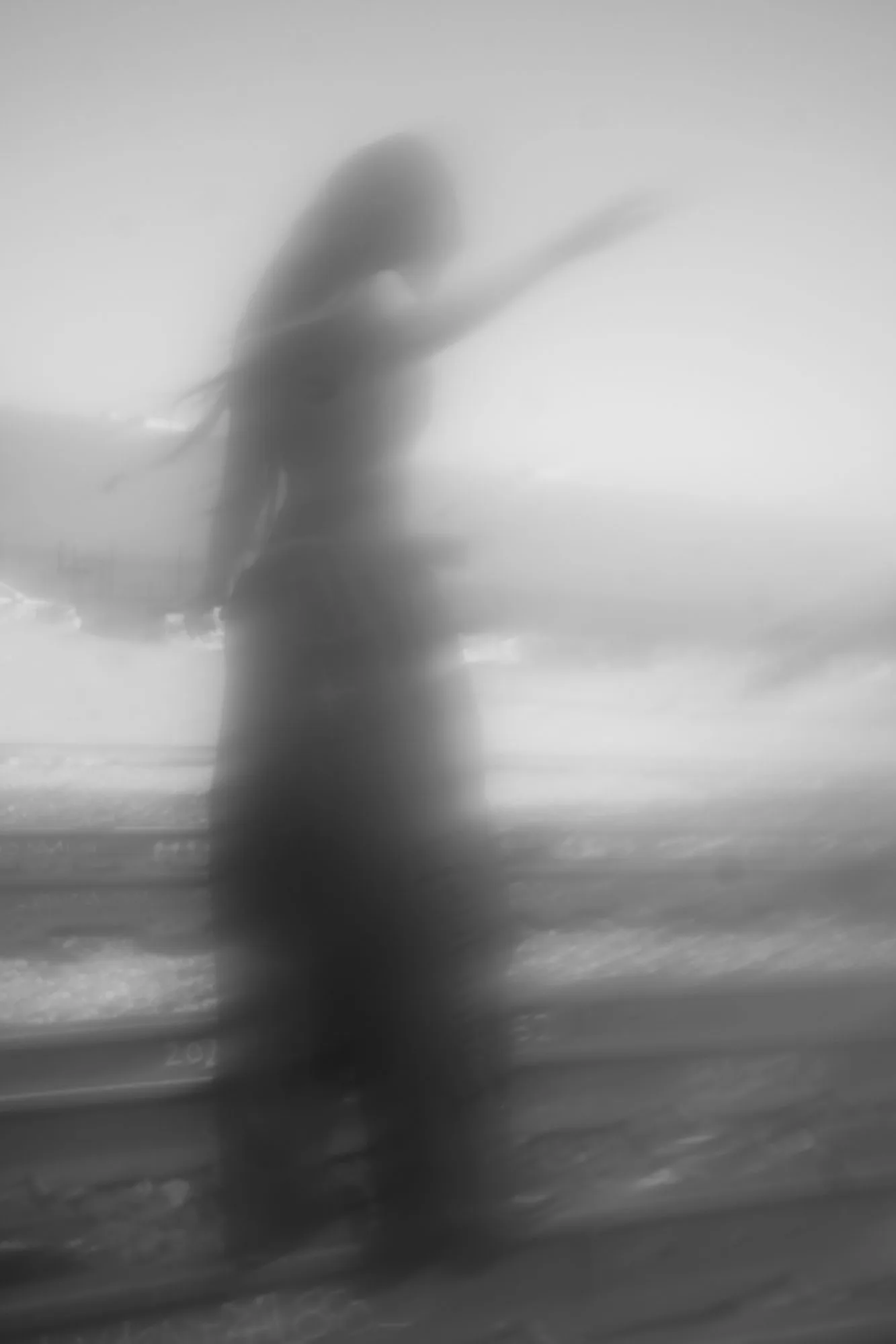 Blurred black-and-white long exposure of a figure raising an arm by the sea