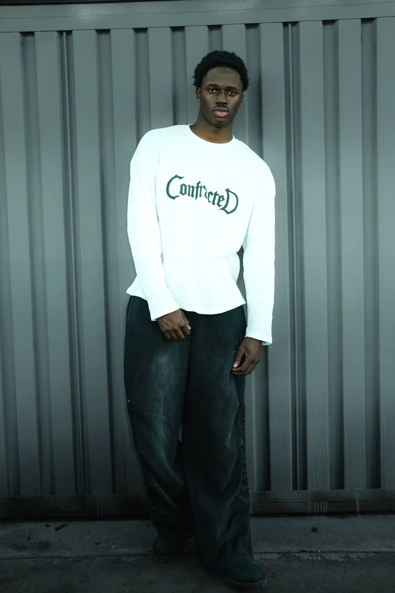 Model in a white Confracted long-sleeve and dark denim standing against a corrugated metal wall