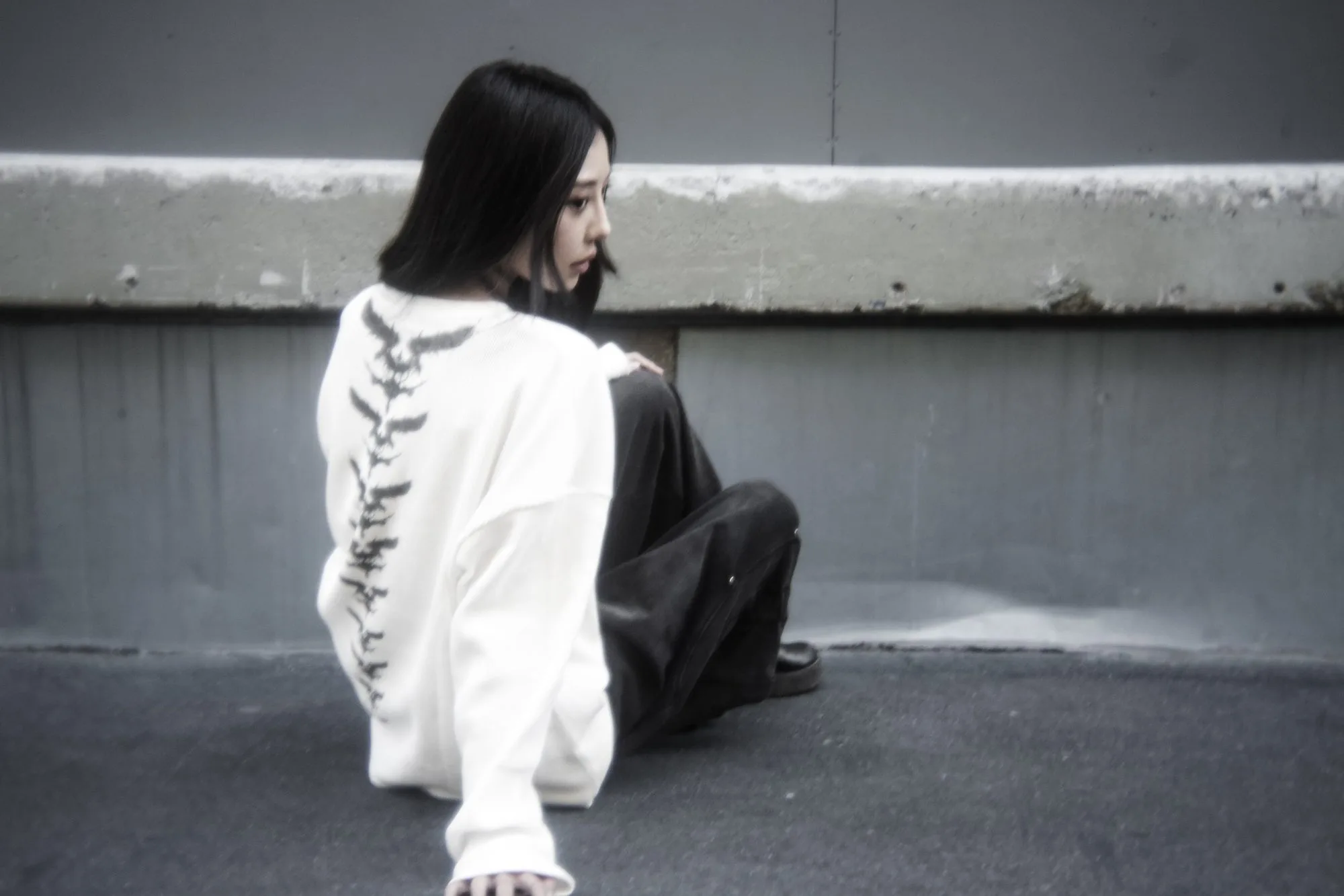 Model seated on asphalt in a white Confracted crewneck with lettering down the spine, looking away