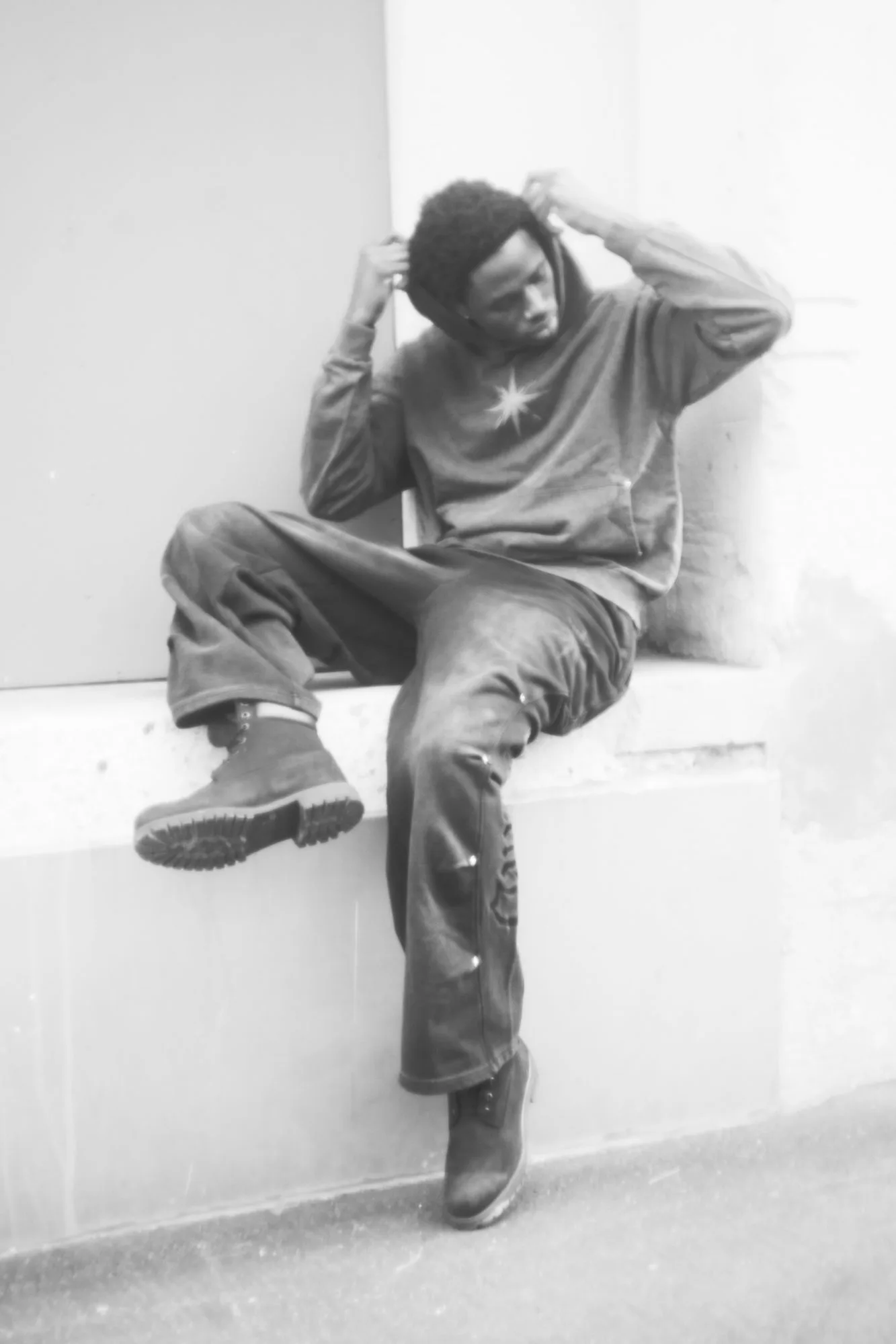 Black-and-white portrait of a model in a hoodie and flared denim seated on a ledge, pulling at the hood