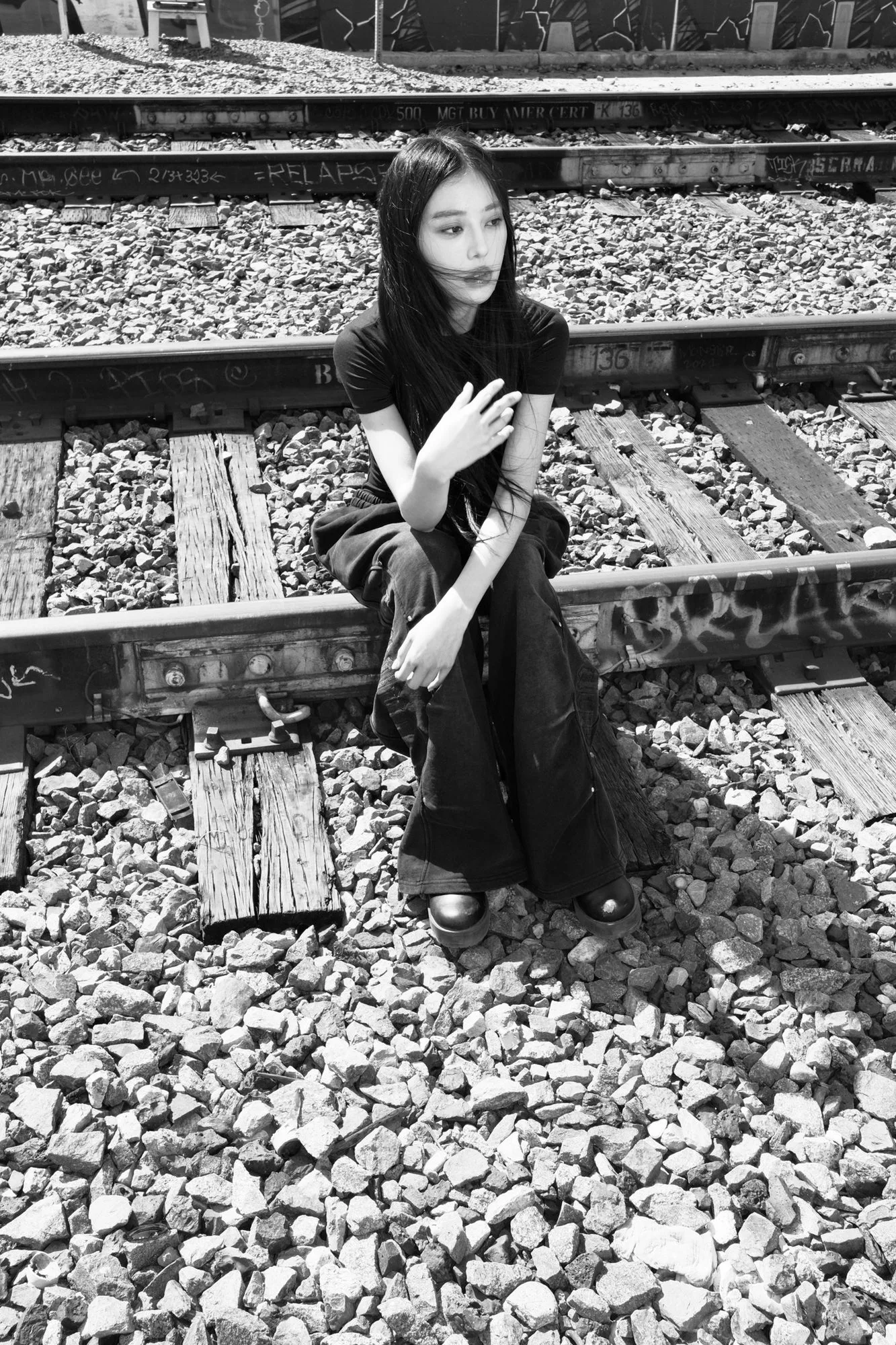 Black-and-white photo of a model seated on graffiti-marked railroad tracks in flared trousers