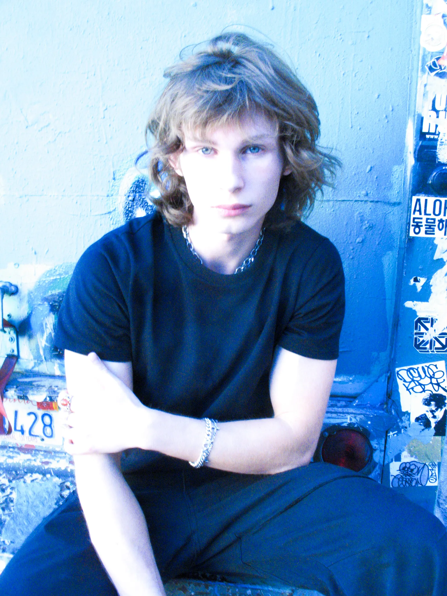 Blue-toned portrait of Dylan Wayne in a chain necklace in front of a sticker-covered wall