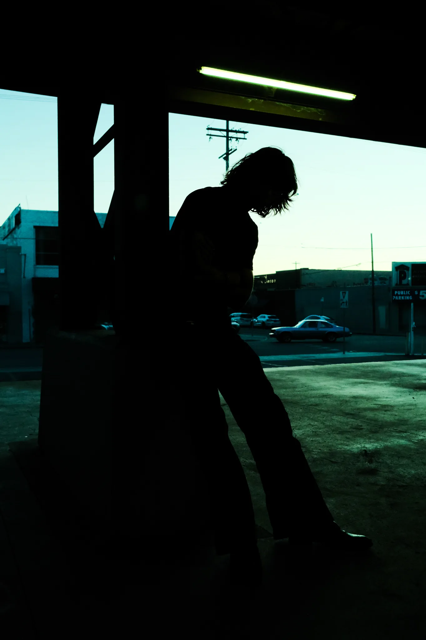Silhouette of Dylan Wayne leaning against a column beneath a fluorescent tube at dusk