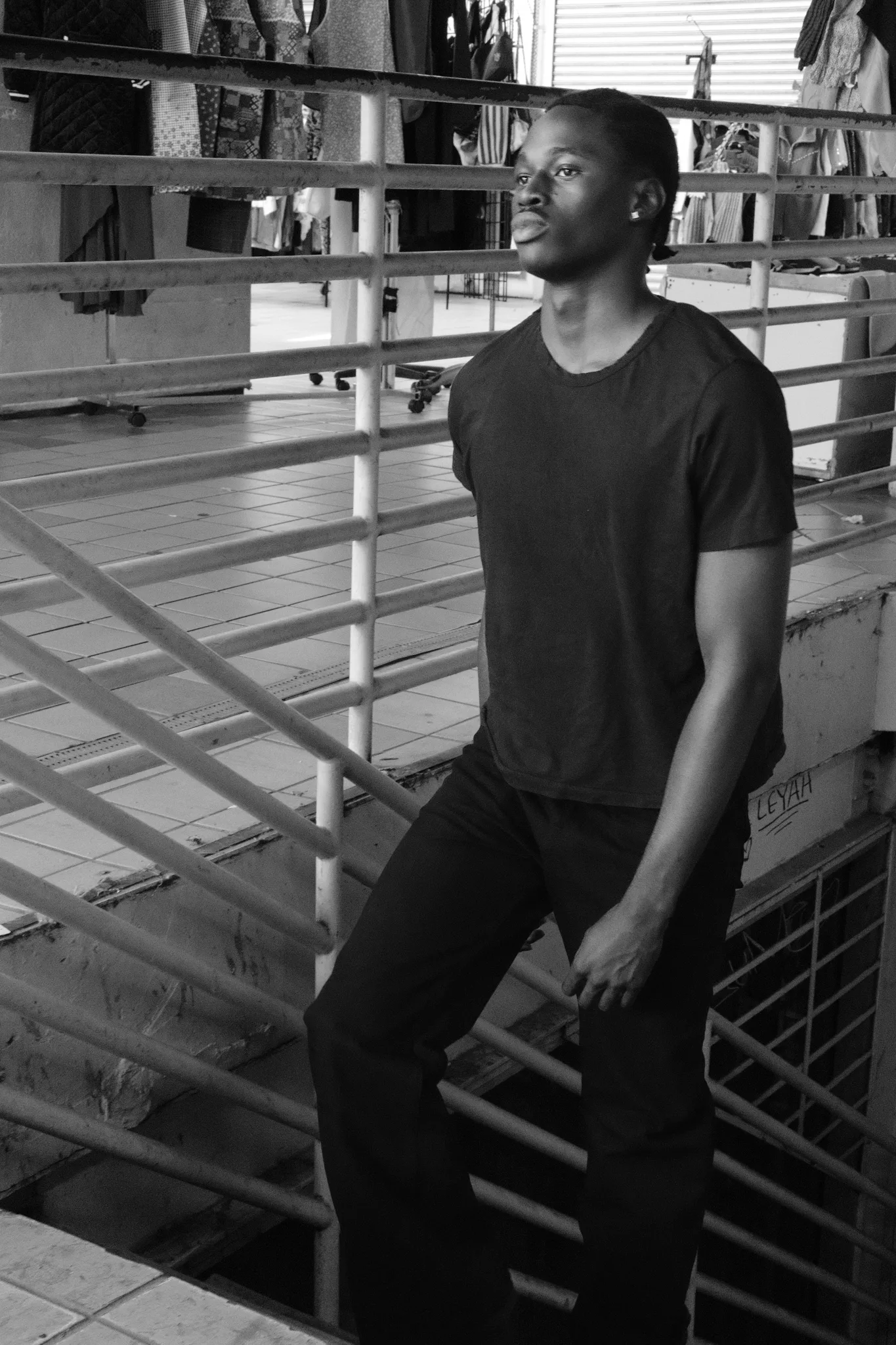 Black-and-white portrait of a model standing at white railings in a garment market, clothing racks behind
