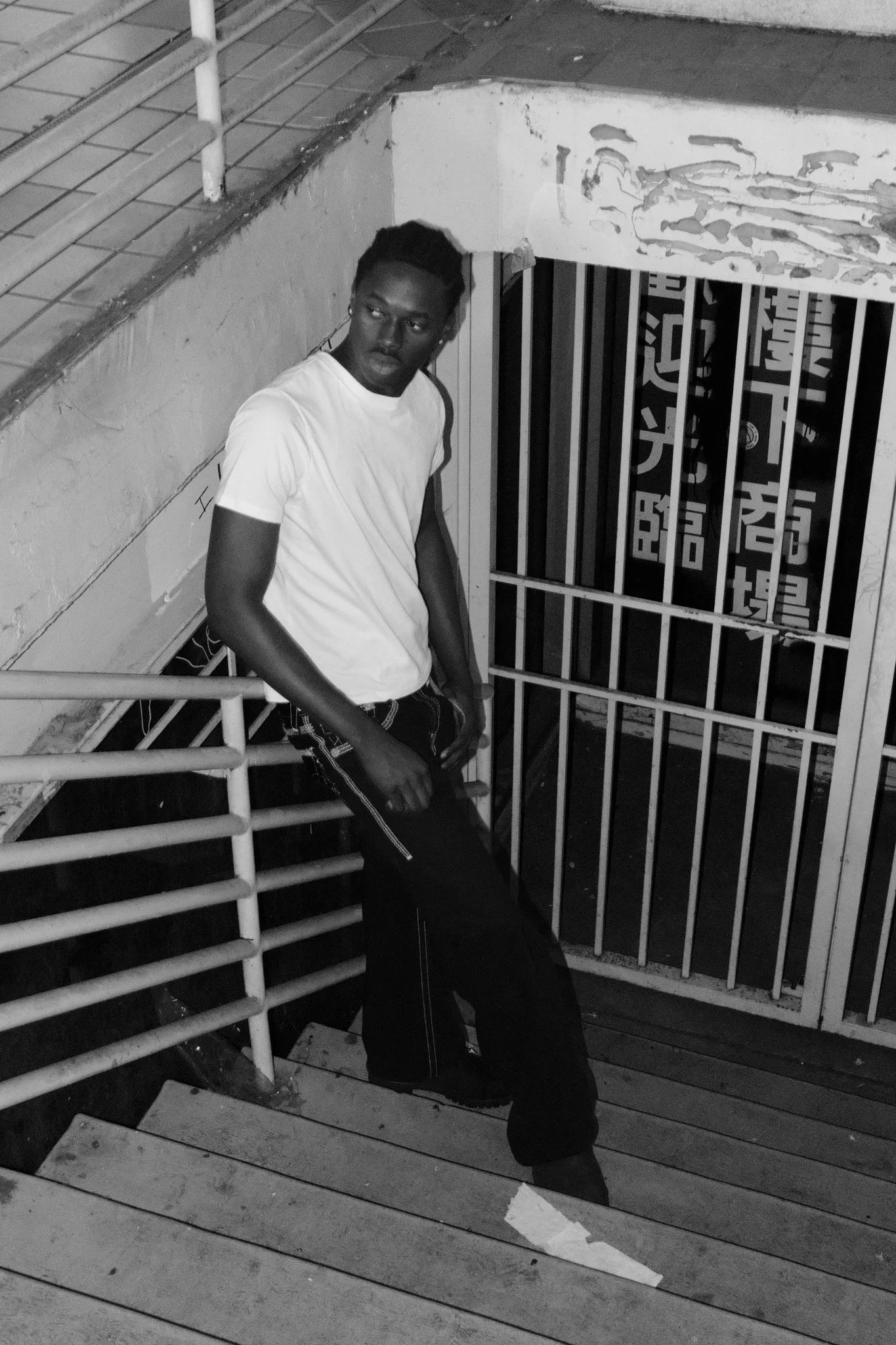 Black-and-white portrait of a model in a white tee on stairs beside shuttered storefronts with Chinese signage