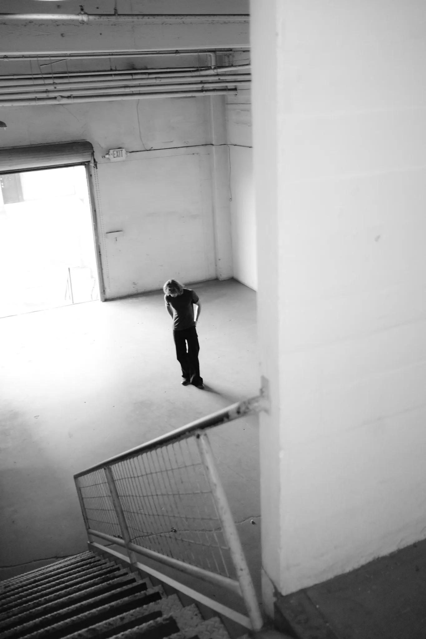 Black-and-white view from a stairwell of a figure crossing an empty warehouse toward a bright loading door