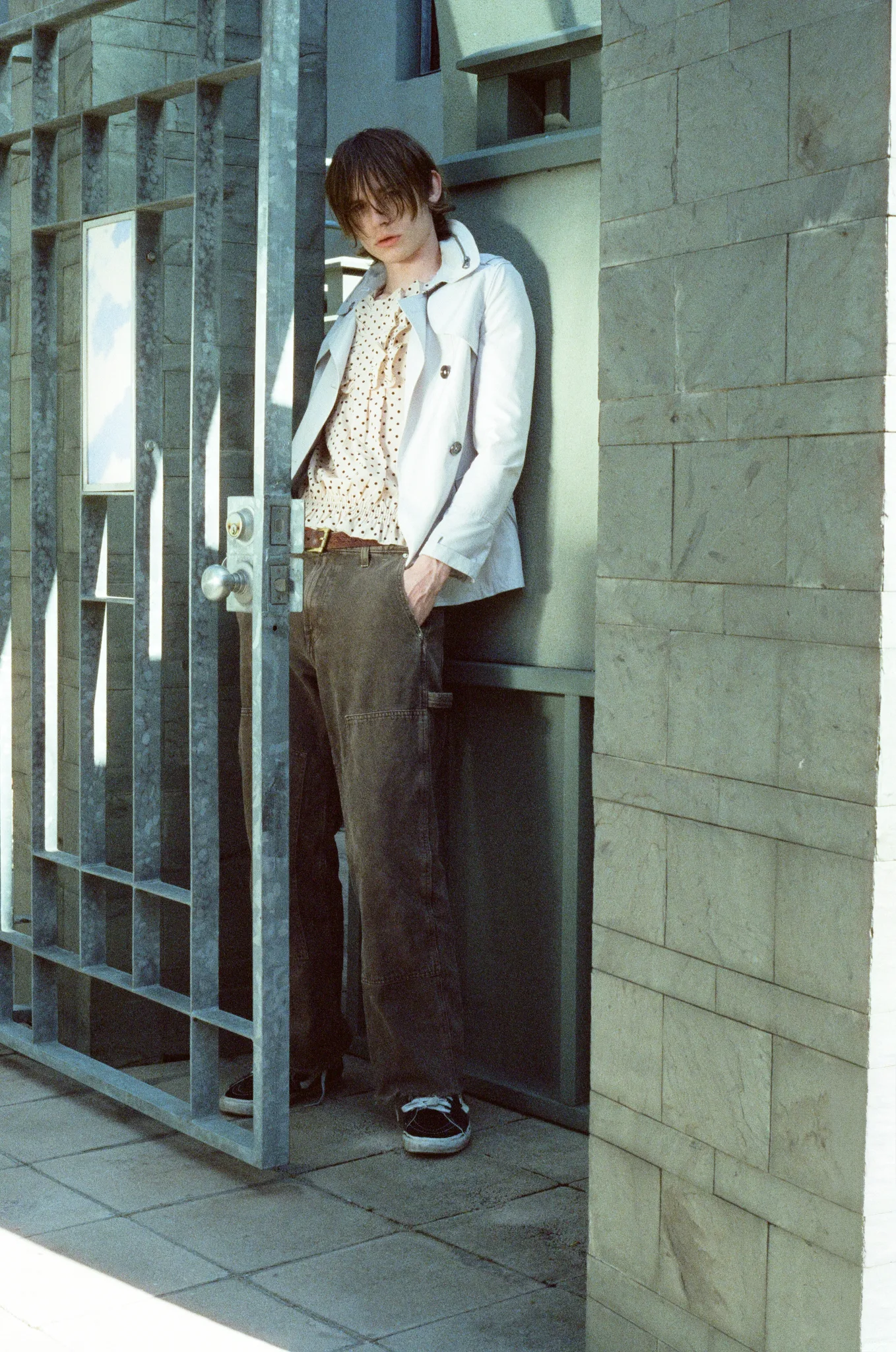 Model in a cream jacket and floral-print shirt leaning in a doorway behind an open metal gate