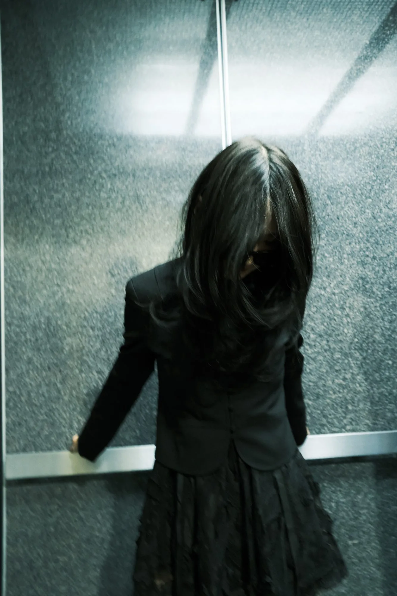 Model with long hair covering her face in a steel elevator, wearing a black blazer and ruffled skirt
