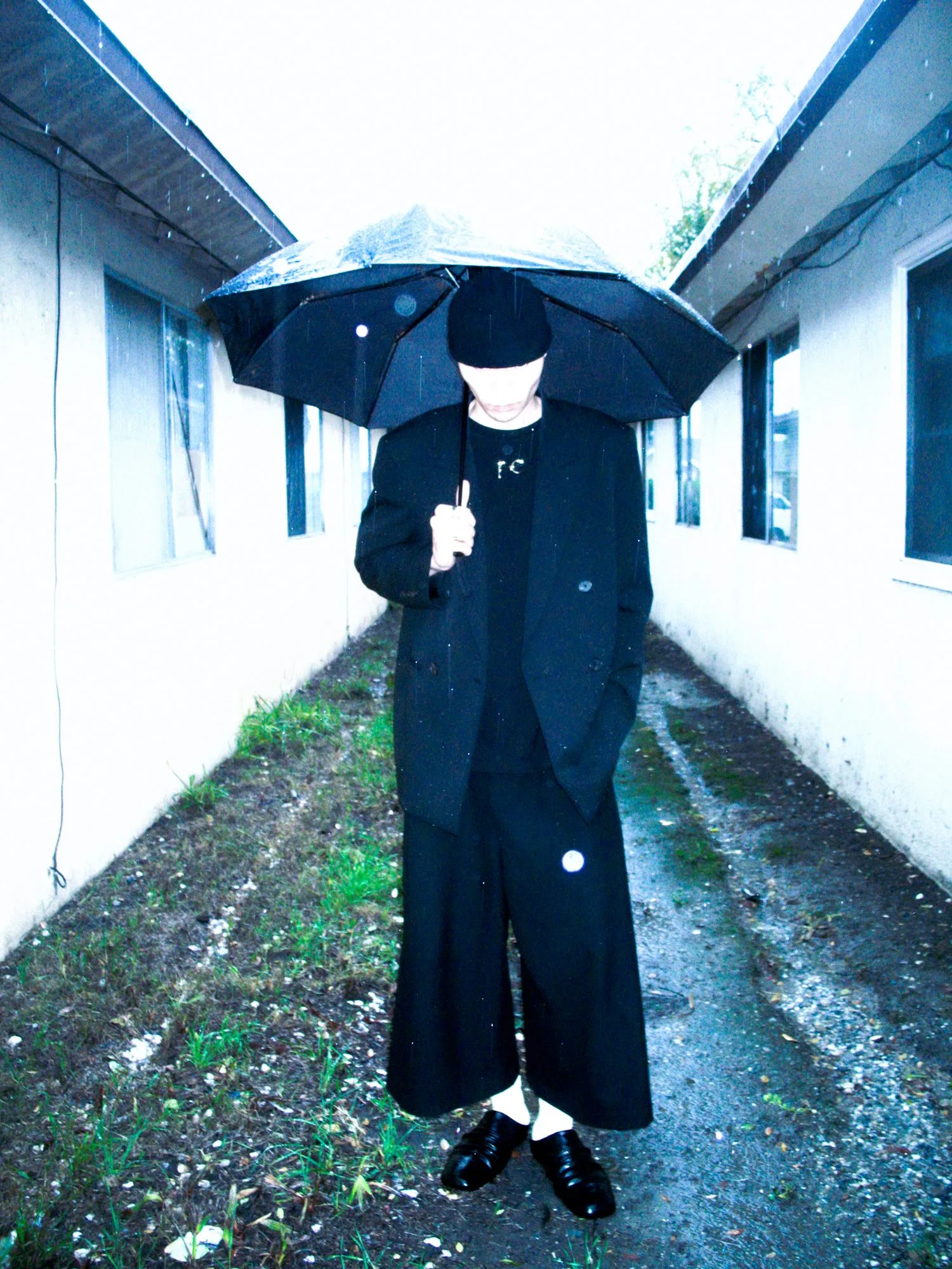 Model in oversized black tailoring holding an umbrella in the rain, standing in an alley between white buildings