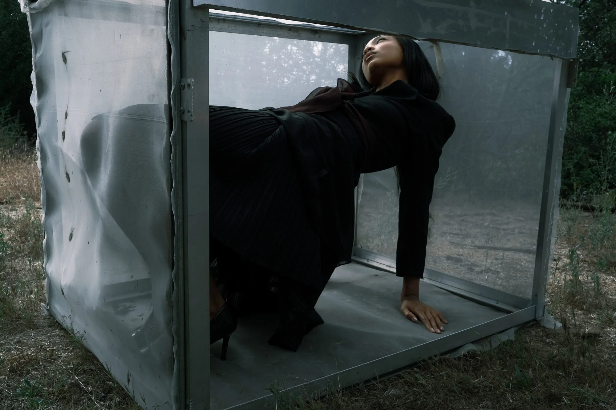 Model in black arching backward inside a translucent mesh cube in a dry field