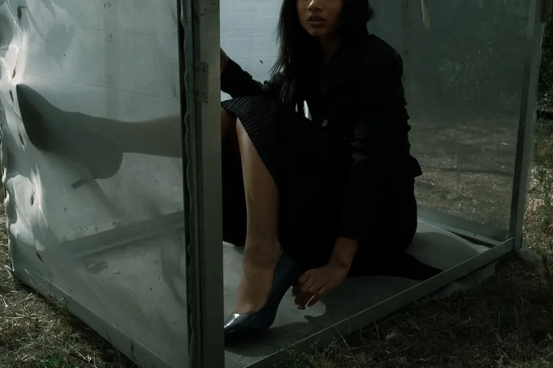 Model in dark clothing seated inside a translucent mesh cube, adjusting her heel
