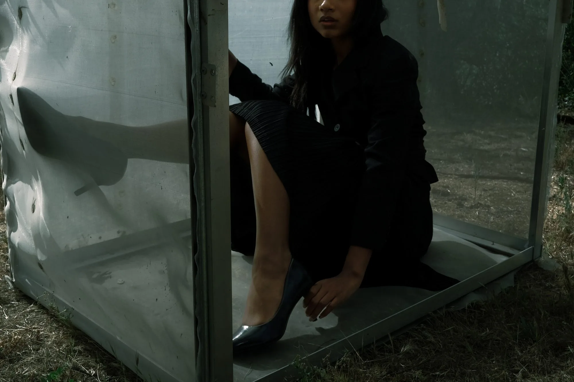 Model in dark clothing seated inside a translucent mesh cube, adjusting her heel