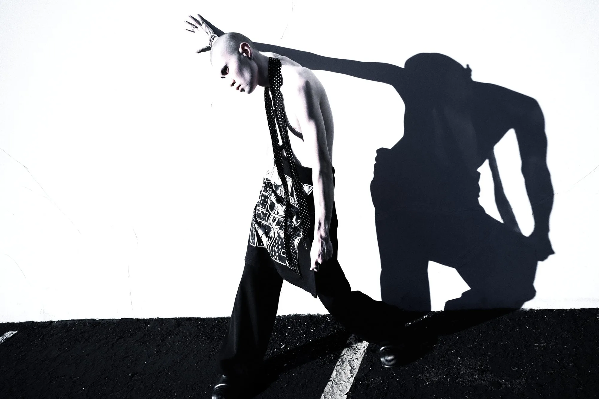 Black-and-white editorial of a model with spiked hair leaning forward, casting a hard kneeling shadow on the wall behind