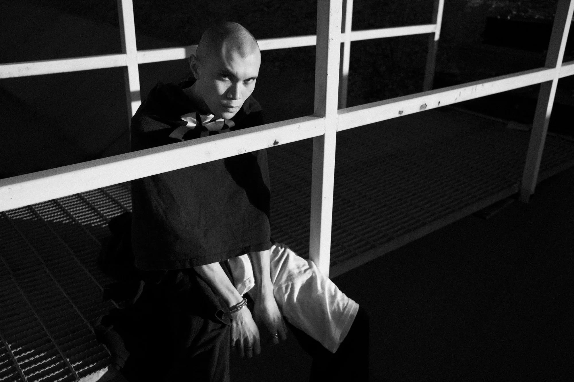 Black-and-white portrait of a model crouched beneath white metal railings at night