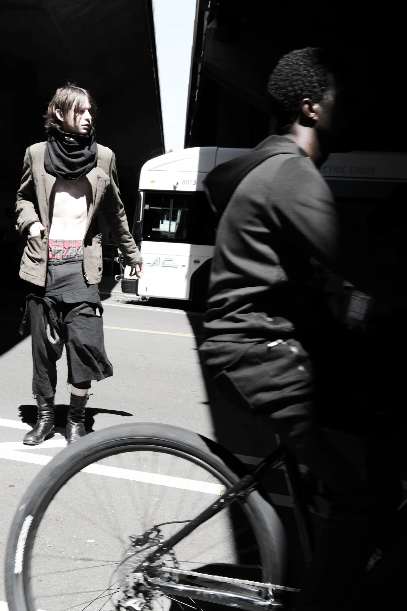 High-contrast street frame of a model in a scarf and open shirt as a cyclist rolls past a city bus