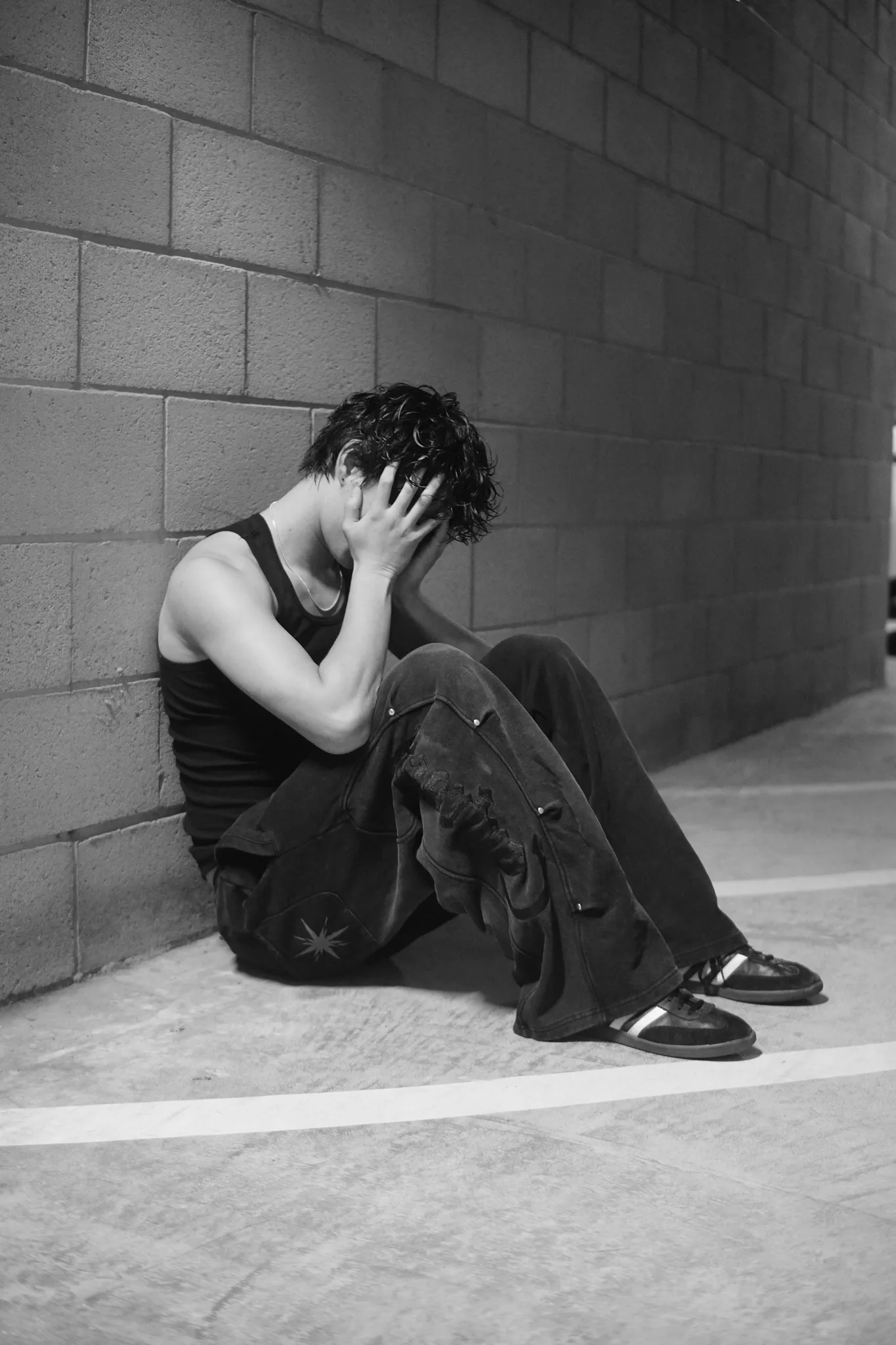 Black-and-white photo of a model seated against a cinder-block wall, hands pulled through his hair