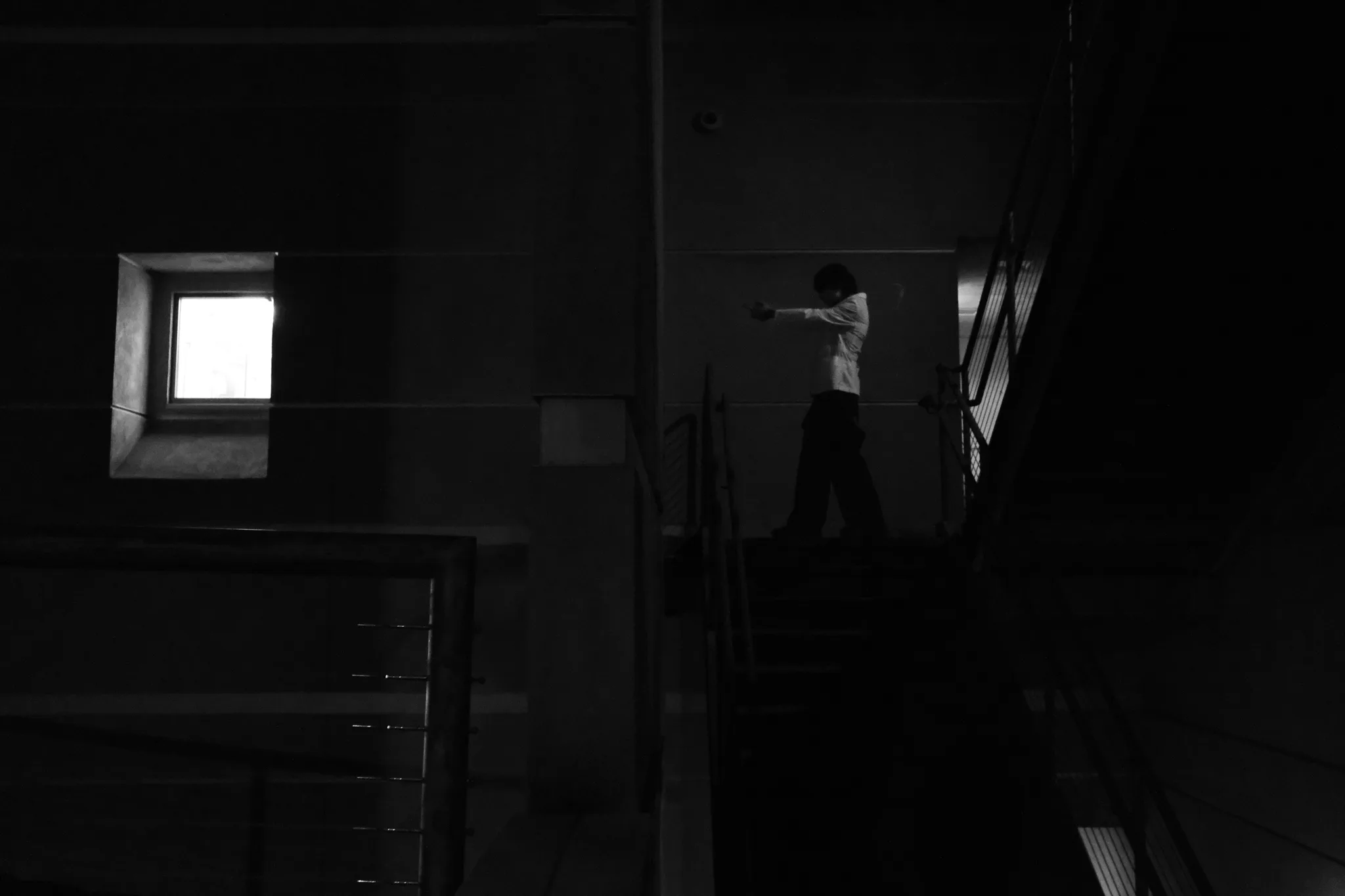 Black-and-white wide shot of a figure in a white shirt on a dark stairwell, lit by a single window
