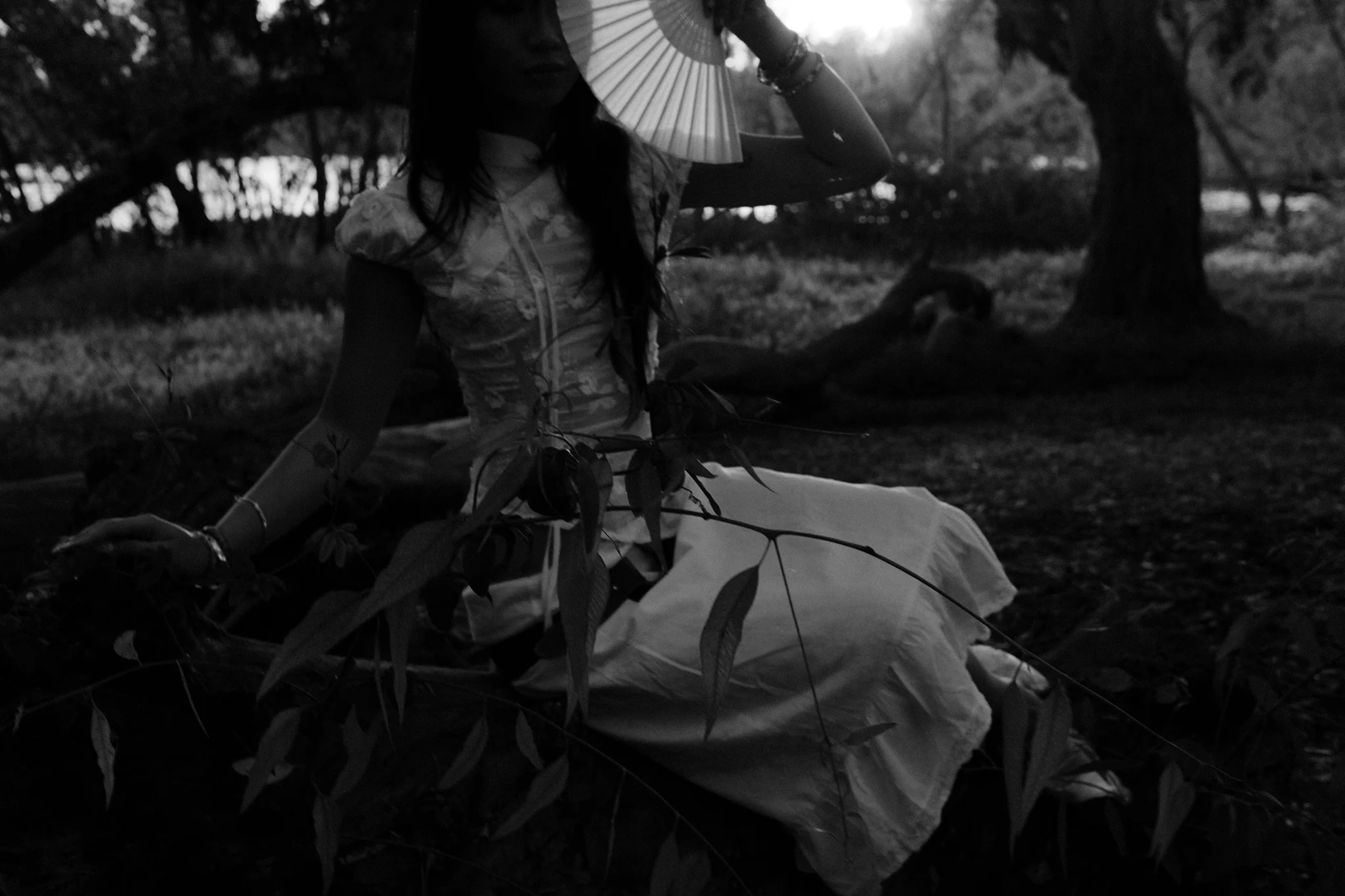 Black-and-white photo of a model in a white lace dress seated among branches, holding a paper fan