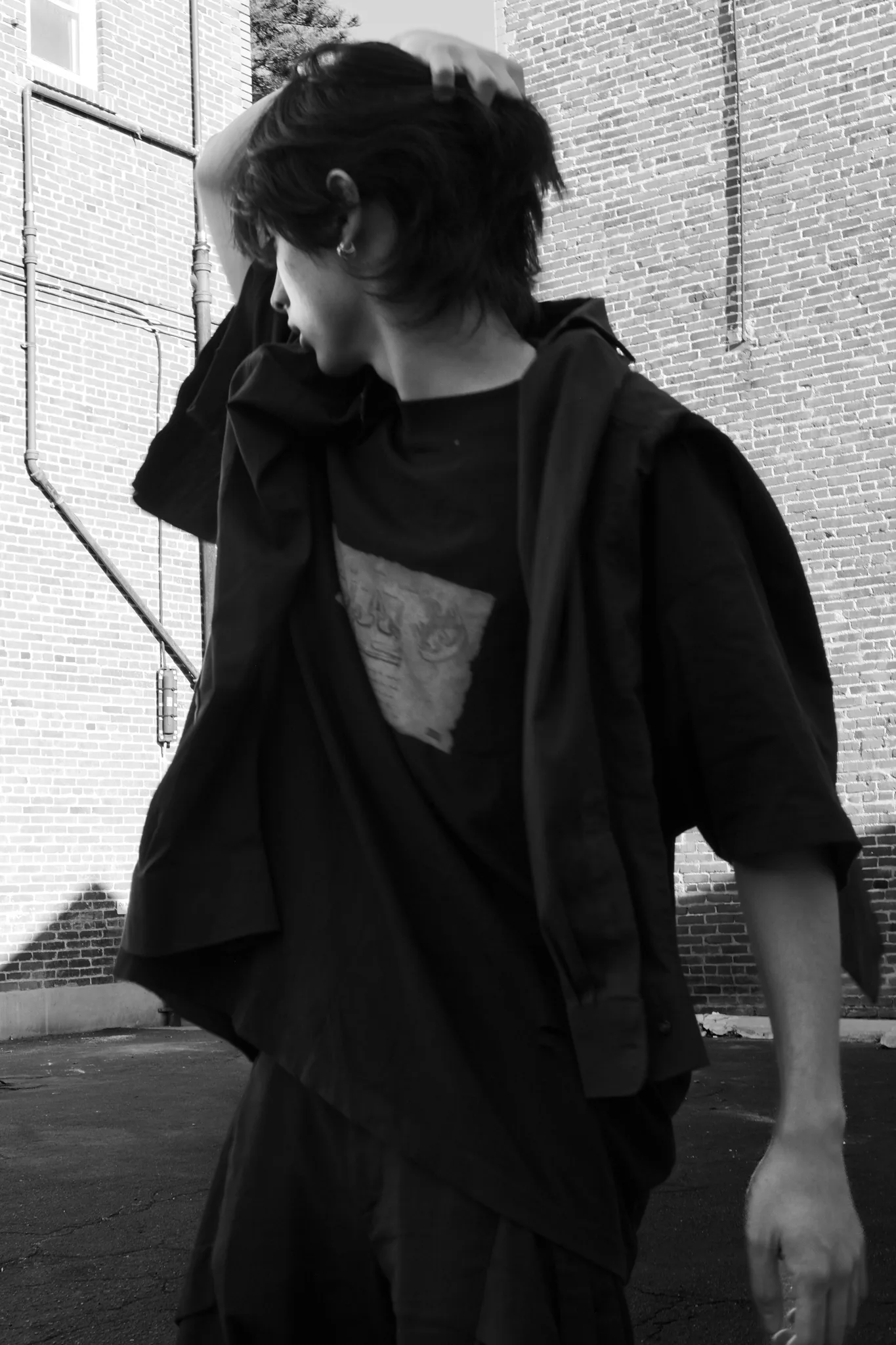 Black-and-white frame of a model in layered black clothing between brick buildings, hand raised to his head