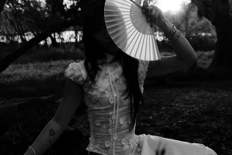 Black-and-white portrait of a model raising a paper fan against the sun, light flaring through its ribs